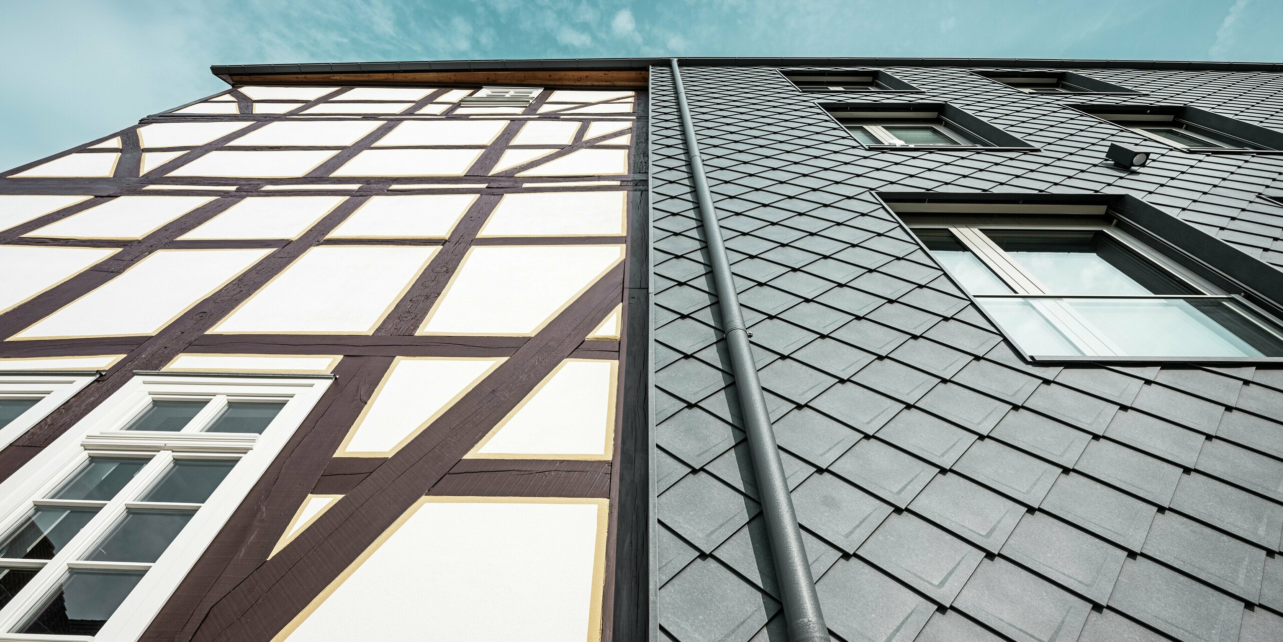 Nahaufnahme der Fassade des Volkshauses in Homberg, DE, mit modernen PREFA Wandrauten 29 × 29 in P.10 Steingrau und historischem Fachwerk. Die Aluminiumverkleidung vereint zeitlose Eleganz, Witterungsschutz und Langlebigkeit, harmonisch ergänzt durch eine integrierte Dachentwässerung. Die funktionelle Entwässerung des Regenwassers erfolgt über die PREFA Kastenrinne mit anschließendem Ablaufrohr. 