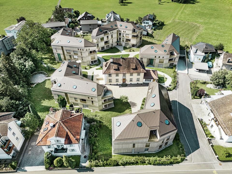 Vista aerea del moderno complesso residenziale di Liebwylen, in Svizzera.  Gli edifici sono progettati con tetti in alluminio PREFALZ di colore marrone e sono dotati di lucernari rotondi. Al centro si trova la villa storica, circondata da nuovi edifici disposti in modo armonioso e inseriti in un paesaggio verde.