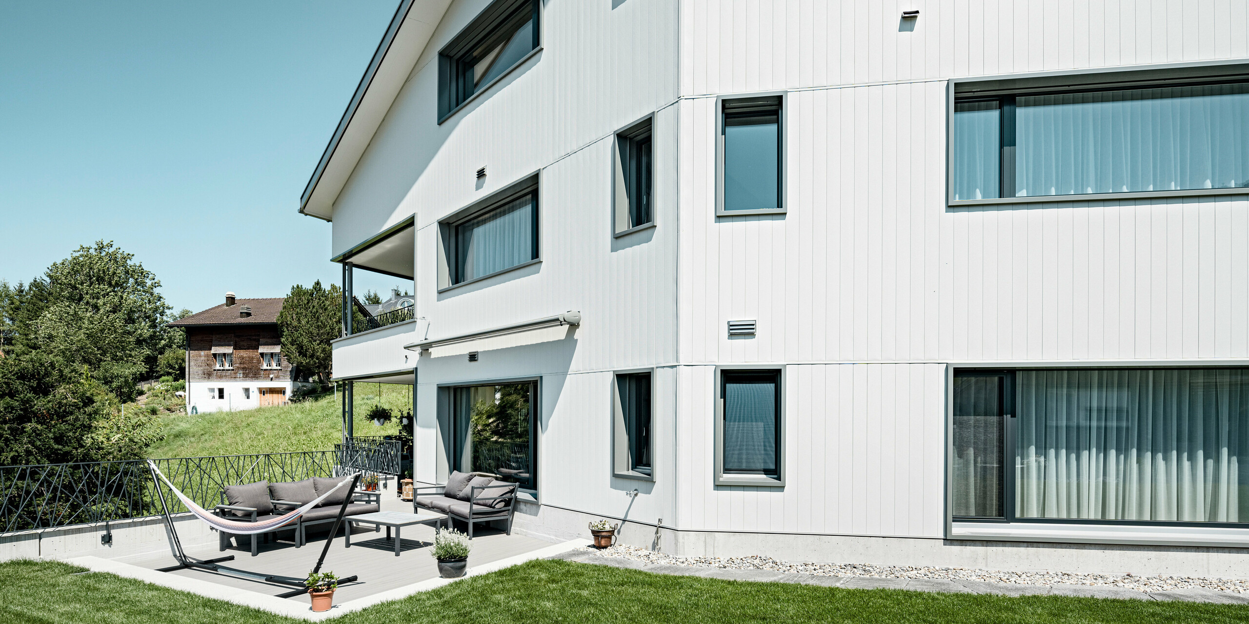 Moderne Wohnkultur in Weissbad, Schweiz: Das Mehrfamilienhaus besticht durch seine PREFA Siding Aluminiumfassade in P.10 Prefaweiß. Die vertikale Anordnung der robusten Fassadenpaneele ergänzt die klare, minimalistische Struktur des Gebäudes. Die Fassade bildet einen stilvollen Kontrast zum gepflegten Grün des Gartens und der einladenden Terrasse, was das Haus zu einem perfekten Beispiel für die Kombination von PREFA Design und Wohnkomfort macht.