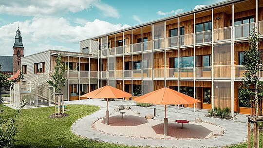 Die Rückseite der Kindertagesstätte in Alzenau zeigt den geschützten Innenhof mit angrenzendem Spielbereich. Zwei orangefarbene Sonnenschirme setzen farbliche Akzente über dem runden Sandspielplatz. Im Hintergrund erstreckt sich der Wohntrakt in nachhaltiger Holzmassivbauweise mit vorgelagerter, filigraner Balkon- und Treppenkonstruktion aus Metall. Links ist die Aluminiumfassade mit PREFA Wandrauten 44 × 44 in Bronze sichtbar – ein robuster und ästhetisch hochwertiger Wetterschutz. Die Kombination aus Aluminium, Holz und Stahl steht sinnbildlich für die intelligente Verbindung von Funktionalität, Nachhaltigkeit und architektonischer Qualität. Der Blick auf die Kirche im Hintergrund verankert das Projekt städtebaulich im gewachsenen Ortsbild von Alzenau.