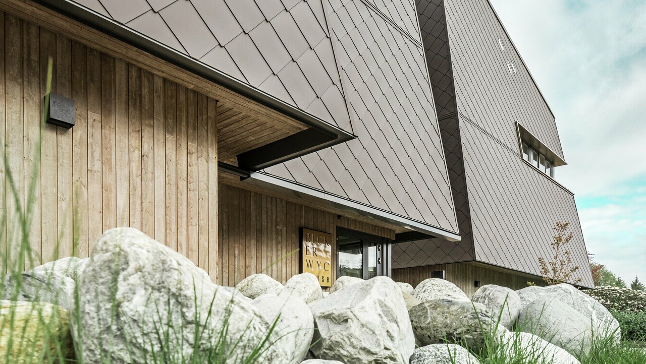 Vista laterale di un hotel, la facciata del piano terra è rivestita verticalmente con legno piuttosto scuro. Sopra c'è una sporgenza della facciata, da qui in poi la facciata è rivestita con pannelli di alluminio color noce.