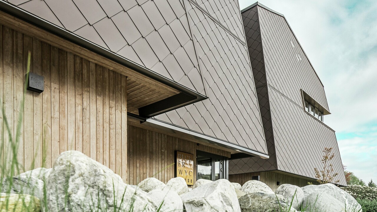 Seitliche Ansicht eines Hotels, die Fassade des Erdgeschosses ist mit eher dunklem Holz vertikal verkleidet. Darüber ist ein Fassadenvorsprung, ab hier ist die Fassade mit Aluminiumplatten in Nussbraun verkleidet.