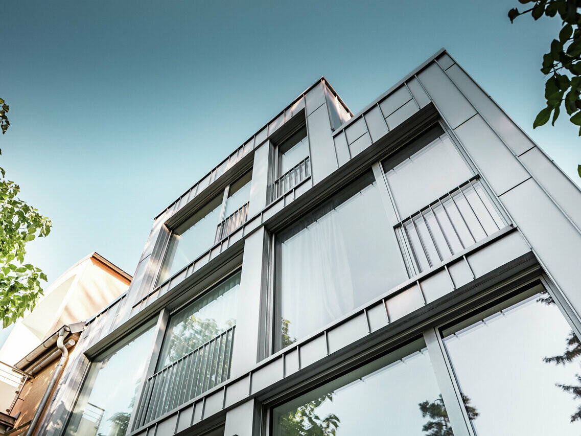 Vista dal basso di un moderno condominio dalla chiara architettura geometrica, caratterizzato da una facciata brillante in alluminio silver metallizzato Prefalz. Le ampie vetrate riflettono il cielo azzurro e la vegetazione circostante. L'edificio è caratterizzato da stretti balconi con ringhiere metalliche che sottolineano il design minimalista.
