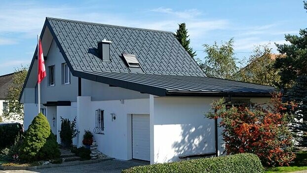 Casa ristrutturata con tetto a due falde e garage adiacente. Il tetto è stato rivestito con tegole PREFA e il garage con Prefalz color antracite. Davanti alla casa è presente un pennone con la bandiera svizzera.