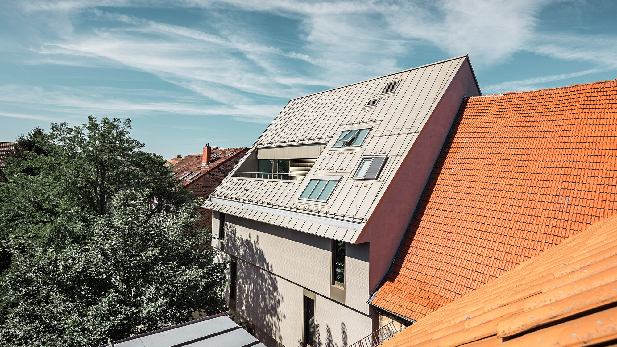 Vista laterale sull'insolita copertura: a sinistra e a destra si ergono degli edifici con tegole piane rosso ruggine, davanti degli alberi.