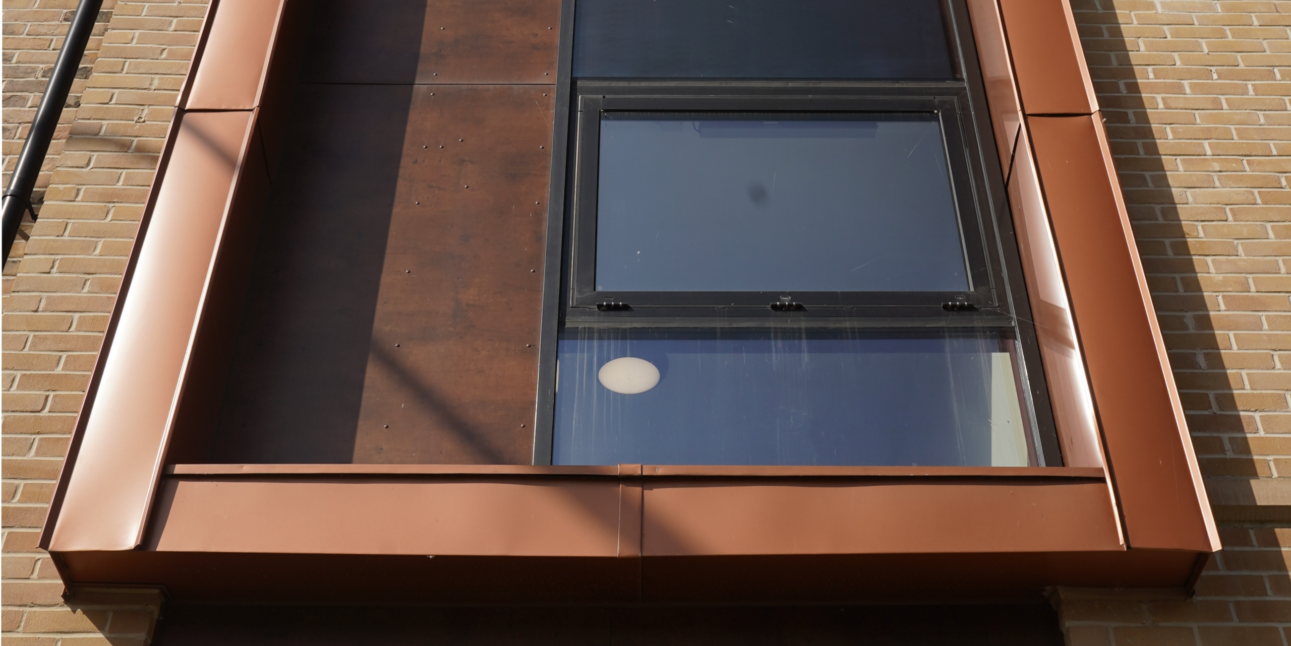 Nahaufnahme eines Fensterbereichs an der Fassade einer Wohnhausanlage in der Nethan Street in Glasgow, umrahmt von FALZONAL in Neukupfer. Dieses qualitativ hochwertige PREFA Aluminiumprodukt ergänzt die klare Glasarchitektur mit seiner warmen Kupfertönung und schafft eine visuelle Verbindung zwischen moderner Bauweise und traditionellen Materialien. Die Verarbeitung von FALZONAL betont die präzise und anspruchsvolle Gestaltung des Wohngebäudes.