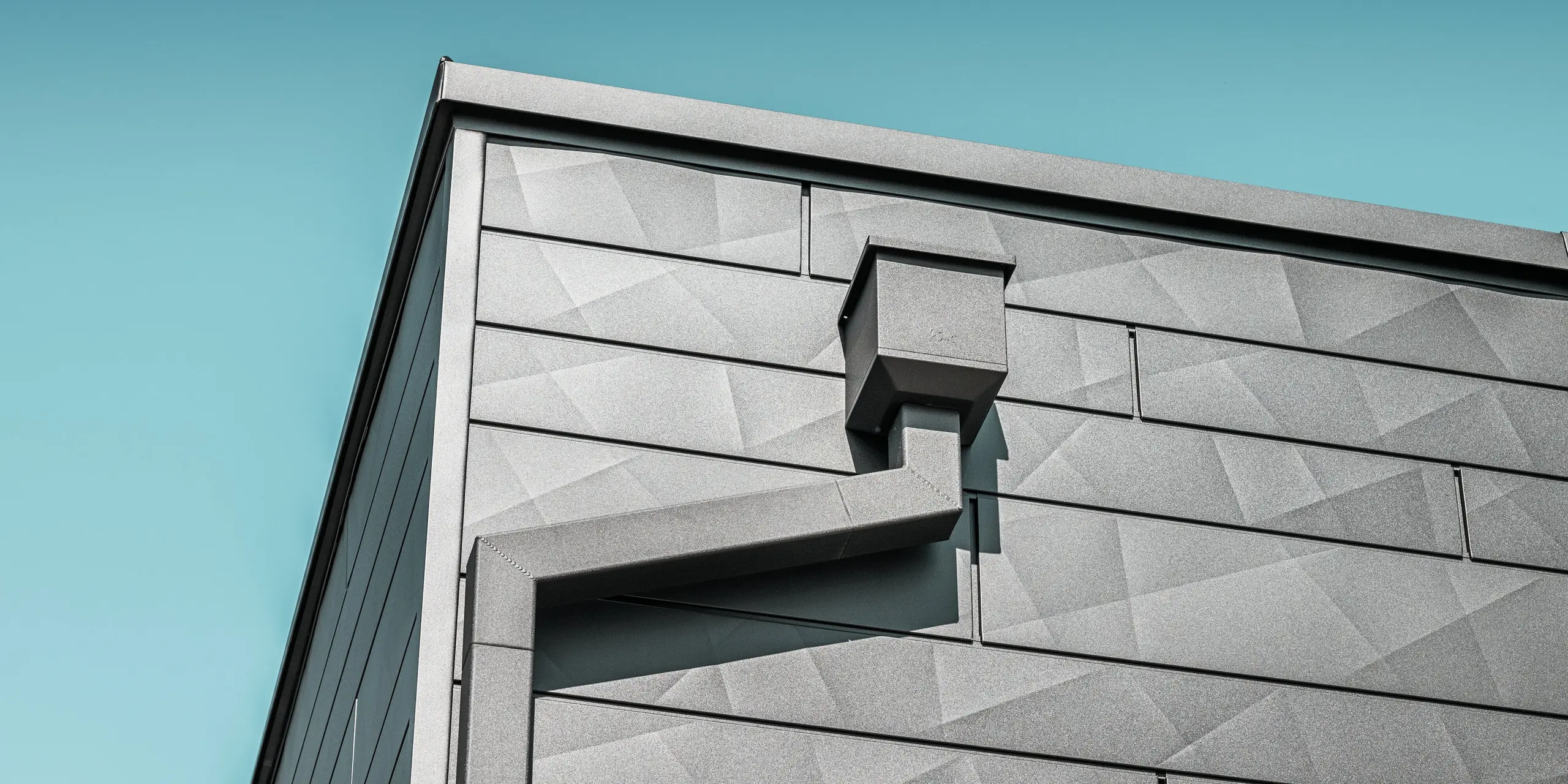 Die Fassade des sanierten Einfamilienhauses in Teuschnitz zieren PREFA Siding.X Aluminiumpaneele in der Farbe P.10 Schwarz mit ihrer einzigartigen, geometrischen Textur. Der Wasserfangkasten und das Quadratrohr von PREFA fügen sich elegant in das Design ein und bieten eine funktionale Lösung für die Entwässerung. Die präzise Verarbeitung und die harmonische Integration der Elemente unterstreichen die ästhetische Qualität und Langlebigkeit der PREFA Aluminiumprodukte.
