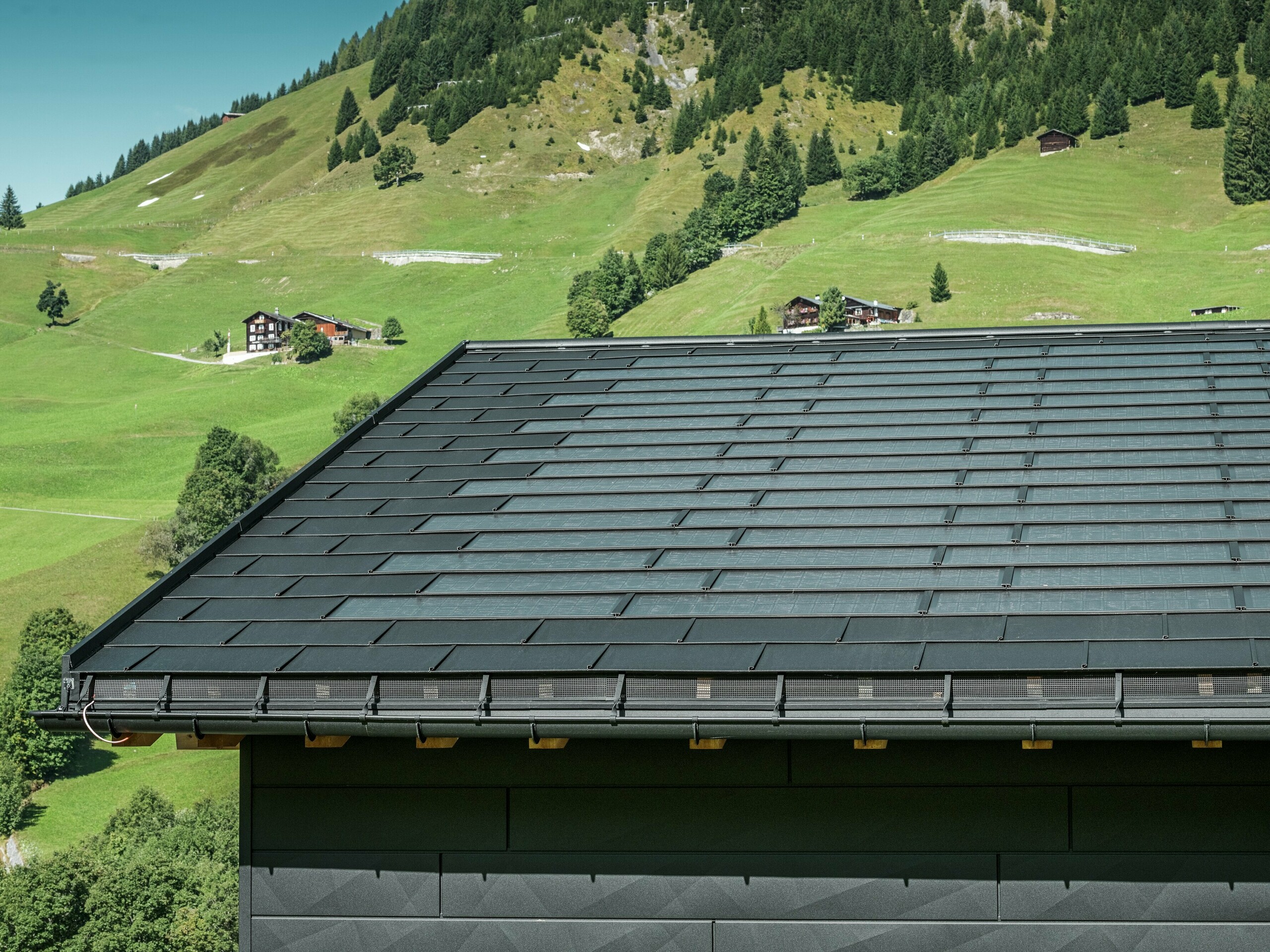 Primo piano della grondaia di una moderna casa unifamiliare nei dintorni alpini di Ascharina, in Svizzera. Il tetto è stato ricoperto con l'innovativa tegola fotovoltaica PREFA e la tradizionale tegola R.16, entrambe in P.10 nero. Lo smaltimento delle acque piovane è garantito dal canale tondo sospeso PREFA e dal tubo pluviale PREFA. Nella parte inferiore dell'immagine sono visibili gli inconfondibili pannelli per facciata doga.X di PREFA. Le linee pulite dei prodotti in alluminio sottolineano il design moderno in mezzo a un pittoresco paesaggio montano.