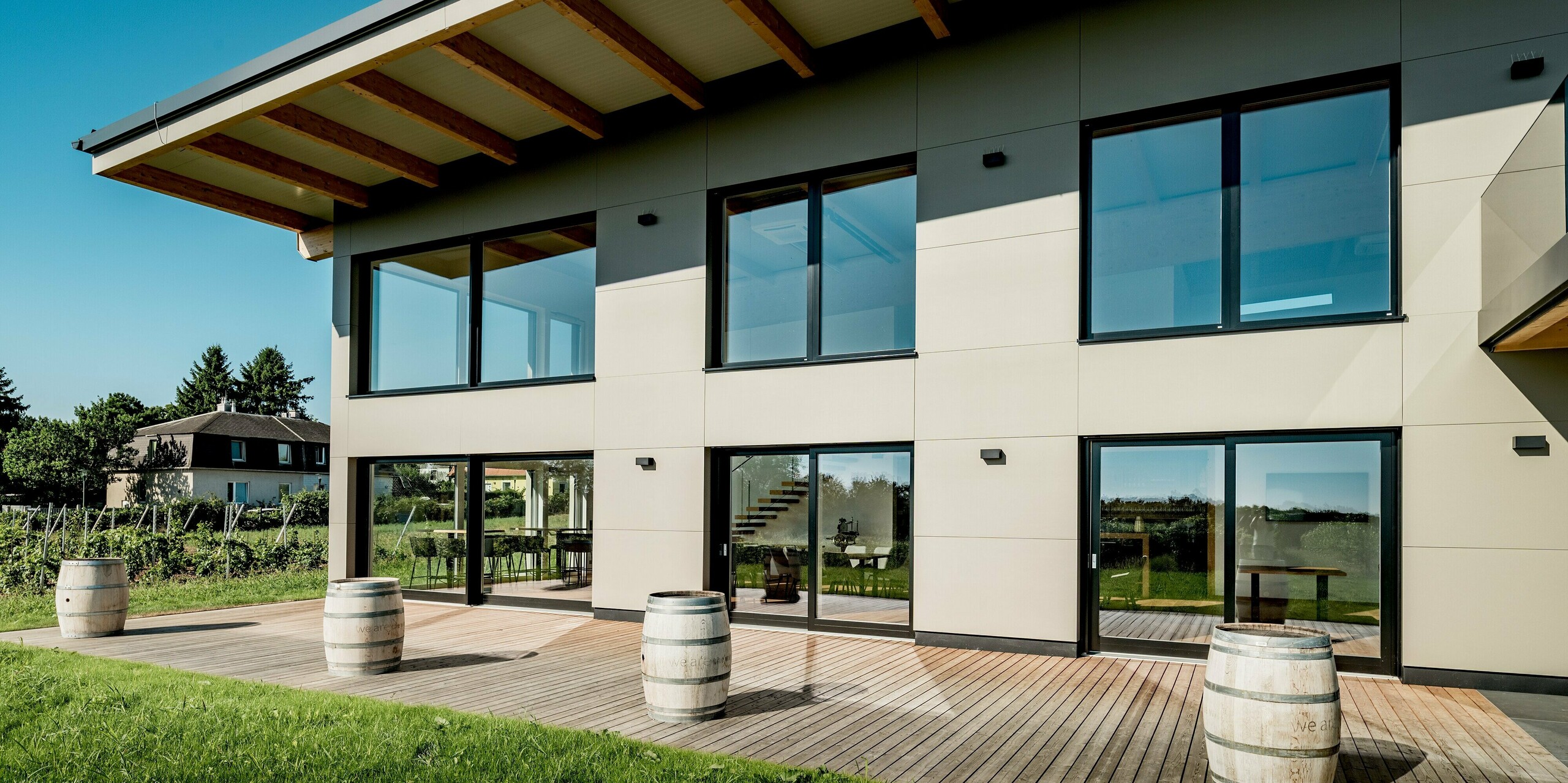 Frontale Ansicht des Weinguts Nigl in Perchtoldsdorf mit moderner Aluminiumfassade von PREFA in P.10 Prefabronze. Die glatte Oberfläche der Verbundplatten und die symmetrische Fensteranordnung prägen das klare, geometrische Fassadenbild. Große Glasflächen reflektieren den Himmel und bieten spannende Einblicke ins lichtdurchflutete Innere mit sichtbarer Treppe. Der darüberliegende Dachvorsprung mit offenen Holzbalken sorgt für natürlichen Sonnenschutz. Im Vordergrund runden Weinbarriquefässer und eine großzügige Holzterrasse die Szene ab. Die PREFA Aluminium Verbundplatte steht hier für moderne Optik, hohe Wetterbeständigkeit und langfristige Farbtreue.