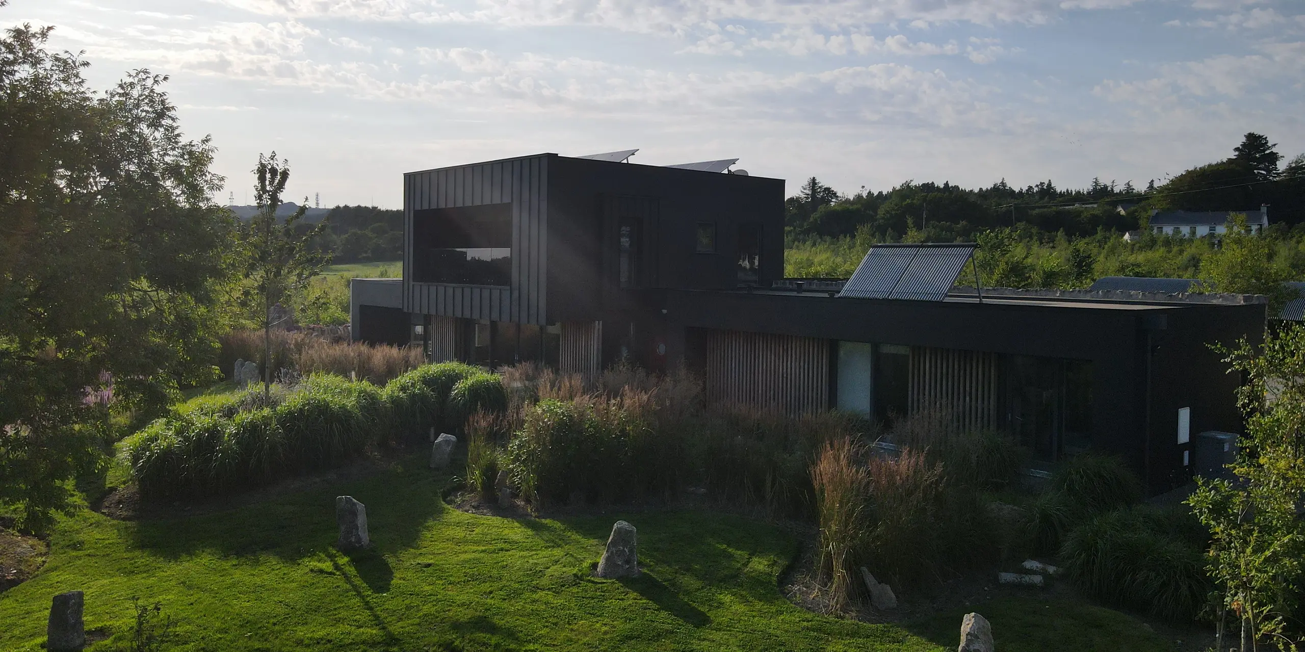 Die Aufnahme zeigt das moderne Einfamilienhaus in Wexford inmitten einer üppigen, wild gestalteten Gartenlandschaft. Besonders ins Auge fällt die schwarze PREFALZ Fassade in P.10 Schwarz, die mit ihrer klaren Linienführung und eleganten Metalloptik einen markanten Kontrast zum satten Grün bildet. Die kraftvolle Abendsonne betont die strukturierten Flächen der Aluminiumverkleidung und unterstreicht ihre hochwertige, witterungsbeständige Qualität. Das architektonische Zusammenspiel aus moderner Kubatur, großen Fensterflächen und robustem PREFA Material macht das Gebäude zu einem eindrucksvollen Highlight.
