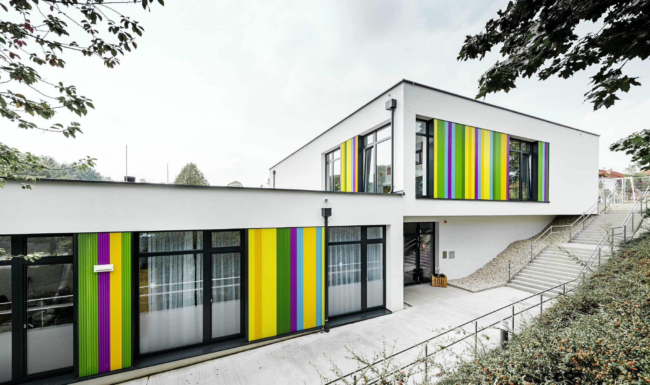 Elemento della facciata colorato di PREFA per la scuola materna ad Hargelsberg. L'edificio ha una copertura piatta e ampie finestre a tutta parete.