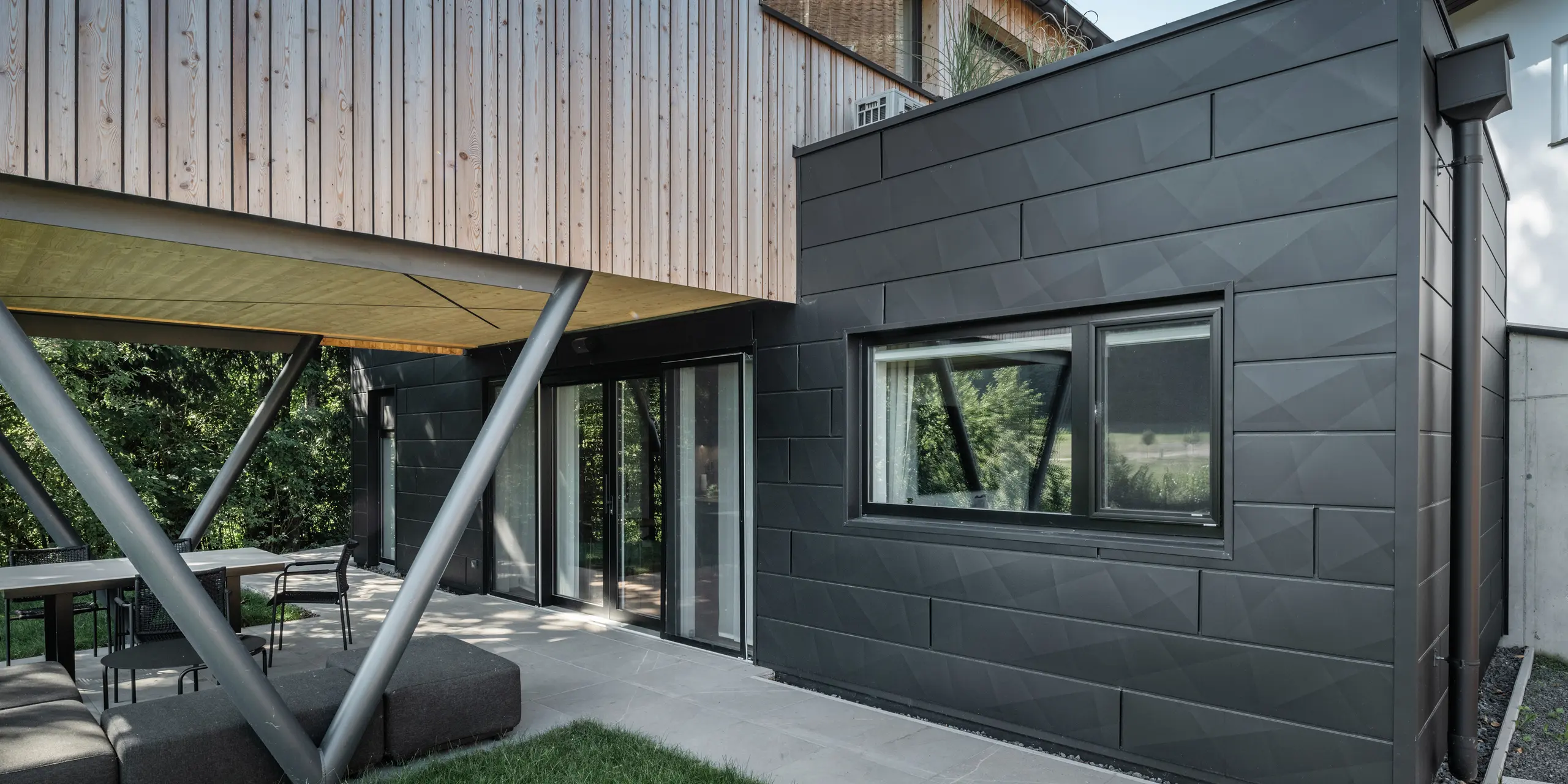 Punto di forza architettonico: “Woodstock Chalet 5” a St. Martin am Innkreis, Alta Austria. La moderna struttura ricettiva per matrimoni è valorizzata dall'uso di pannelli per facciate PREFA Siding.X color antracite. Gli elementi in alluminio creano un forte contrasto con i pannelli in legno chiaro e le strutture di sostegno metalliche. I telai delle finestre scuri si integrano armoniosamente nel design della facciata, conferendo all'edificio un aspetto elegante. Questa immagine mostra l'equilibrio estetico tra la texture naturale del legno e il fascino industriale della facciata in alluminio, che rappresenta il design degli edifici contemporanei.