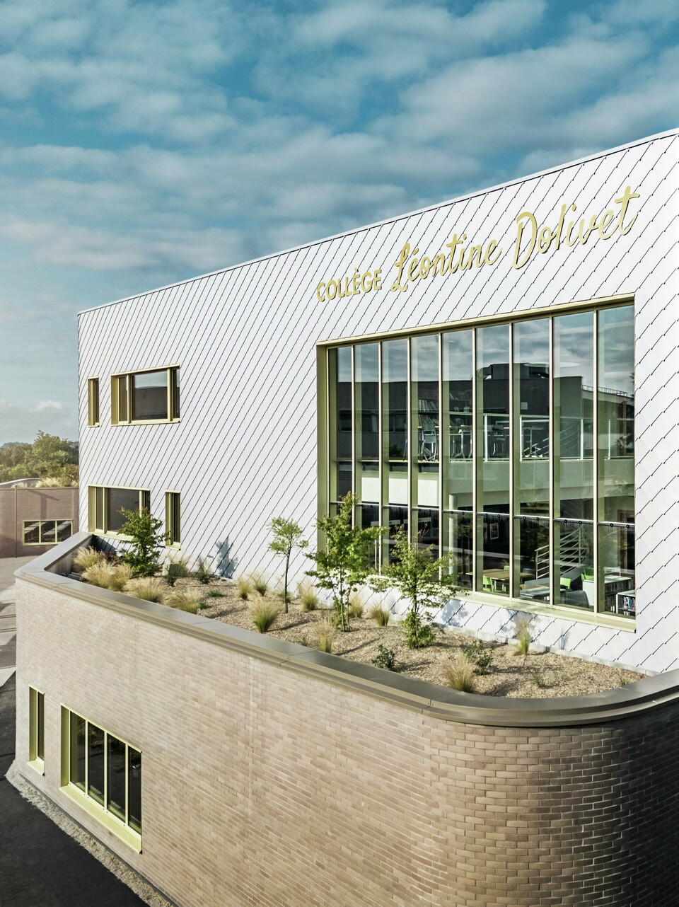 Fotografija prikazuje upečatljiv kut zgrade Collège Léontine Dolivet u Cesson-Sévignéu. Fasada se sastoji od srebrno-metalnih PREFA zidnih rombova, raspoređenih u dijagonalni uzorak koji reflektira svjetlo. Posebno se ističe zakrivljeni oblik gornjeg kata, koji zgradi daje mekani i moderni izgled. Velike staklene površine otvaraju pogled prema unutrašnjosti, dok žuti okviri prozora stvaraju živopisne naglaske. U prvom planu nalazi se zasađeno područje s šljunkom i grmljem, koje arhitekturu skladno nadopunjuje. Lagano oblačno nebo dodatno naglašava metalnu površinu.