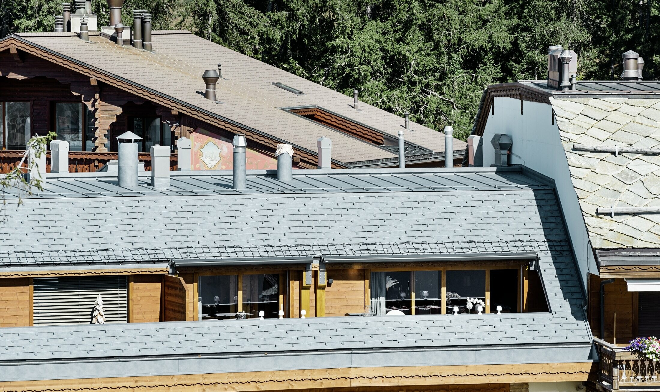 Mehrfamilienhaus in Crans-Montana mit Bergen im Hintergrund und einer Fassade mit verspielten Holzelementen mit einem PREFA Aluminium Dachschindel-Dach in steingrau 