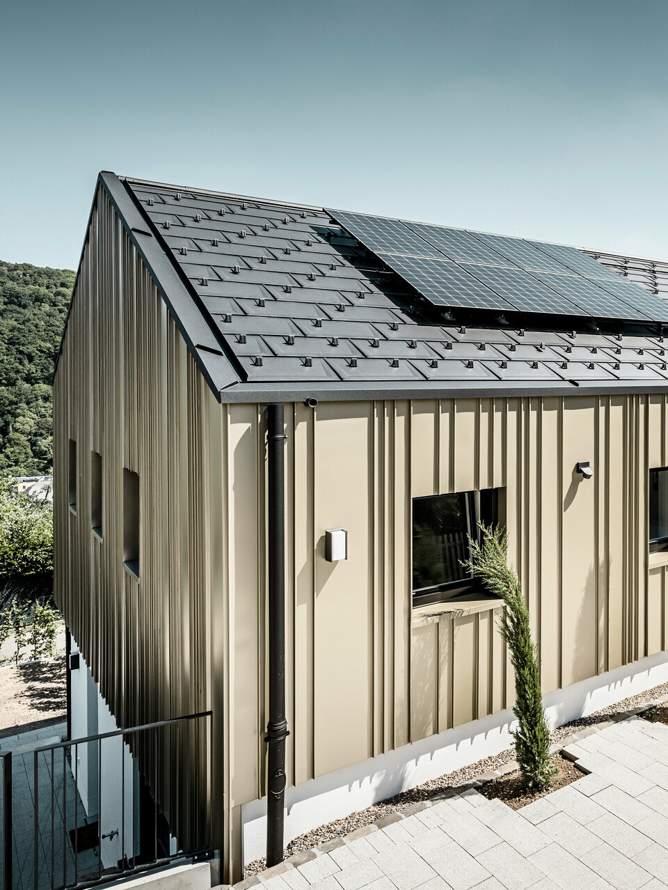 Architektonisch ansprechendes Einfamilienhaus, harmonisch in die Hügellandschaft eingebettet, mit einem hochwertigen PREFA Dachsystem in Form der Dachplatte R.16 in elegantem P.10 Anthrazit. Die Fassade zeichnet sich durch eine geschmackvolle Winkelfalzkonstruktion aus Falzonal in Lichtbronze aus, die das Gebäude mit einem warmen, metallischen Glanz umhüllt. Das Haus verfügt zudem über eine umweltfreundliche Komponente in Form von Solarmodulen, die diskret auf dem Dach integriert sind. Die PV-Anlage sowie die Gebäudehülle aus langlebigem Aluminium betonen das Engagement für nachhaltiges Bauen. Eine minimalistische Garage fügt sich nahtlos an das Hauptgebäude an, während die gepflasterte Einfahrt und sorgfältig platzierte Grünpflanzen das Bild eines modernen, ökologisch bedachten Zuhauses vervollständigen.