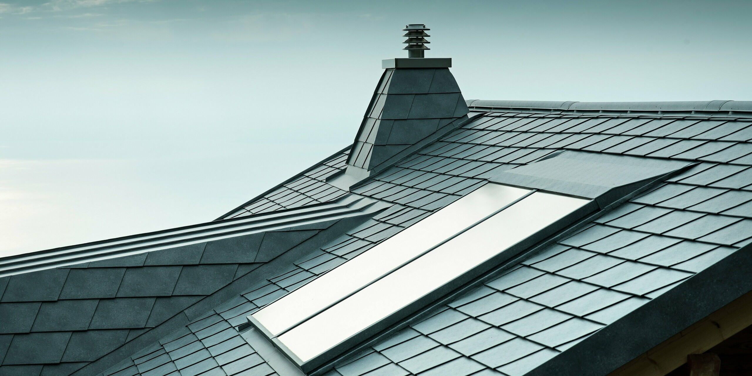 Diese vielseitige Dachansicht zeigt steingraue PREFA-Dachschindeln sowie einen Kamin in Trapezform aus Aluminium. Das langlebige Aluminiumdach ist nicht nur stilvoll, sondern auch äußerst widerstandsfähig. Die exakte Ausführung der Dachgaube und des Dachfensters zeugt von der Detailgenauigkeit der PREFA-Verlegepartner.