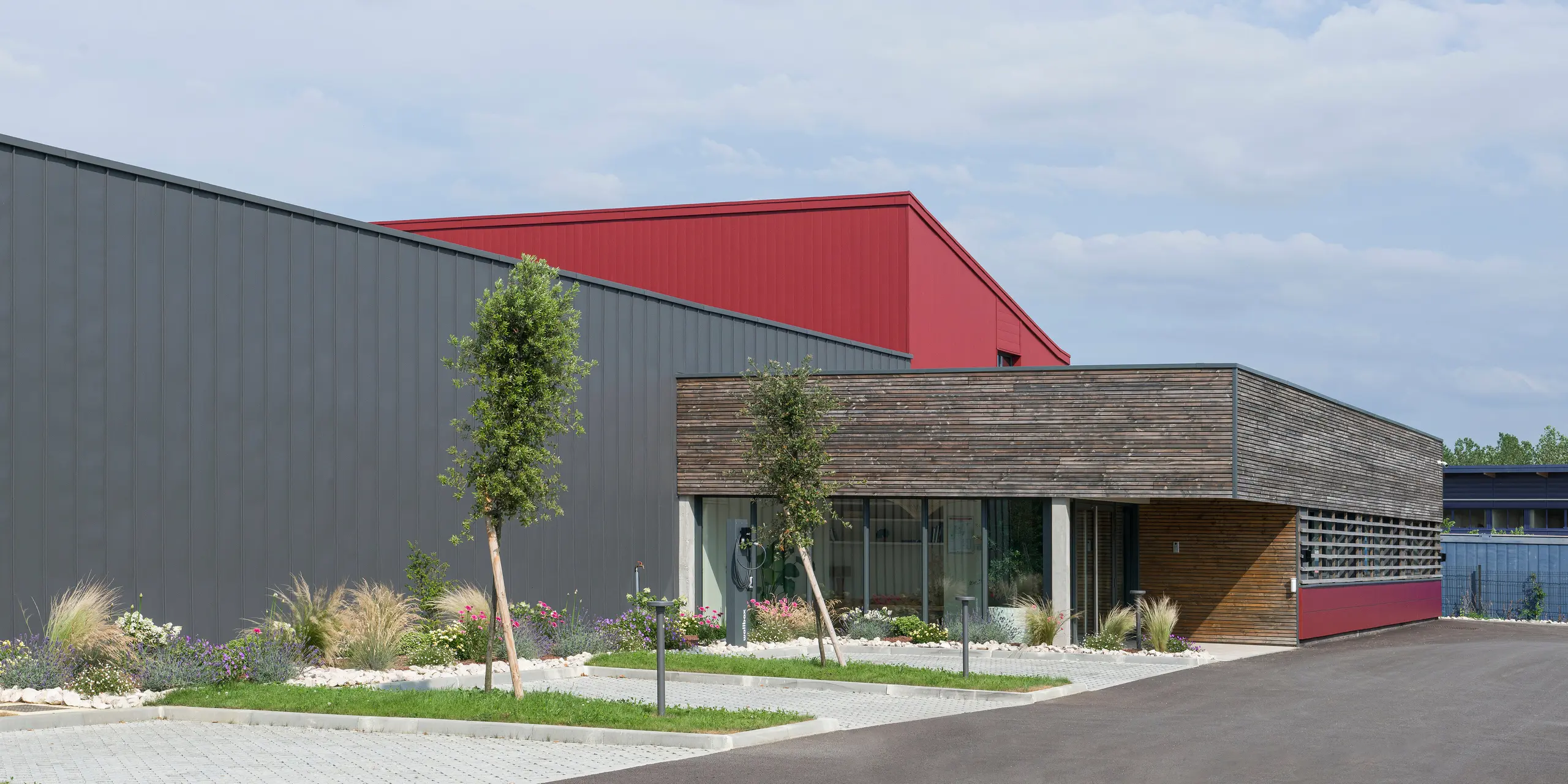 Das Bild zeigt ein modernes Gebäude mit einer auffälligen Kombination aus industriellen und natürlichen Designelementen. Die Fassade kombiniert dunkelgraue und oxydrote Aluminiumelemente, um Dynamik und Textur zu erzeugen. Die oxydroten PREFA Fliesen setzen einen farblichen Akzent. Der untere Teil des Gebäudes mit seiner holzgetäfelten Wand und den großen Fenstern vermittelt eine warme, einladende Atmosphäre, die an einen Büroraum denken lässt. Ein gepflegter Garten vor dem Gebäude vermittelt ein umweltbewusstes und nachhaltiges Image. Die Kombination dieser Elemente spiegelt den Wunsch wider, traditionelle Baupraktiken mit modernen, nachhaltigen Materialien und Designs zu verbinden.