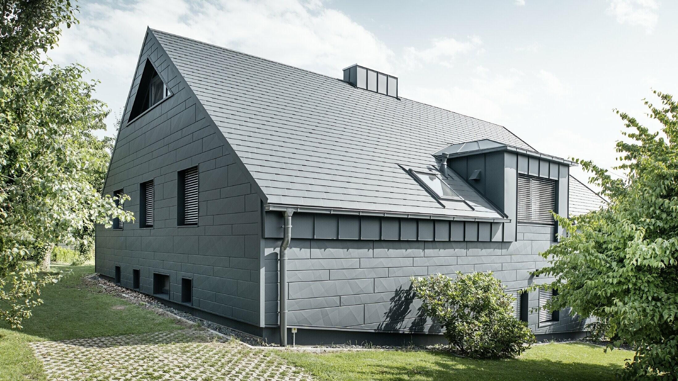 Grande casa unifamiliare con tetto a due falde e molte finestre. L'intera casa è stata rivestita con un involucro in alluminio antracite, sul tetto sono state utilizzate tegole in alluminio PREFA e sulla facciata Doga.X in antracite.