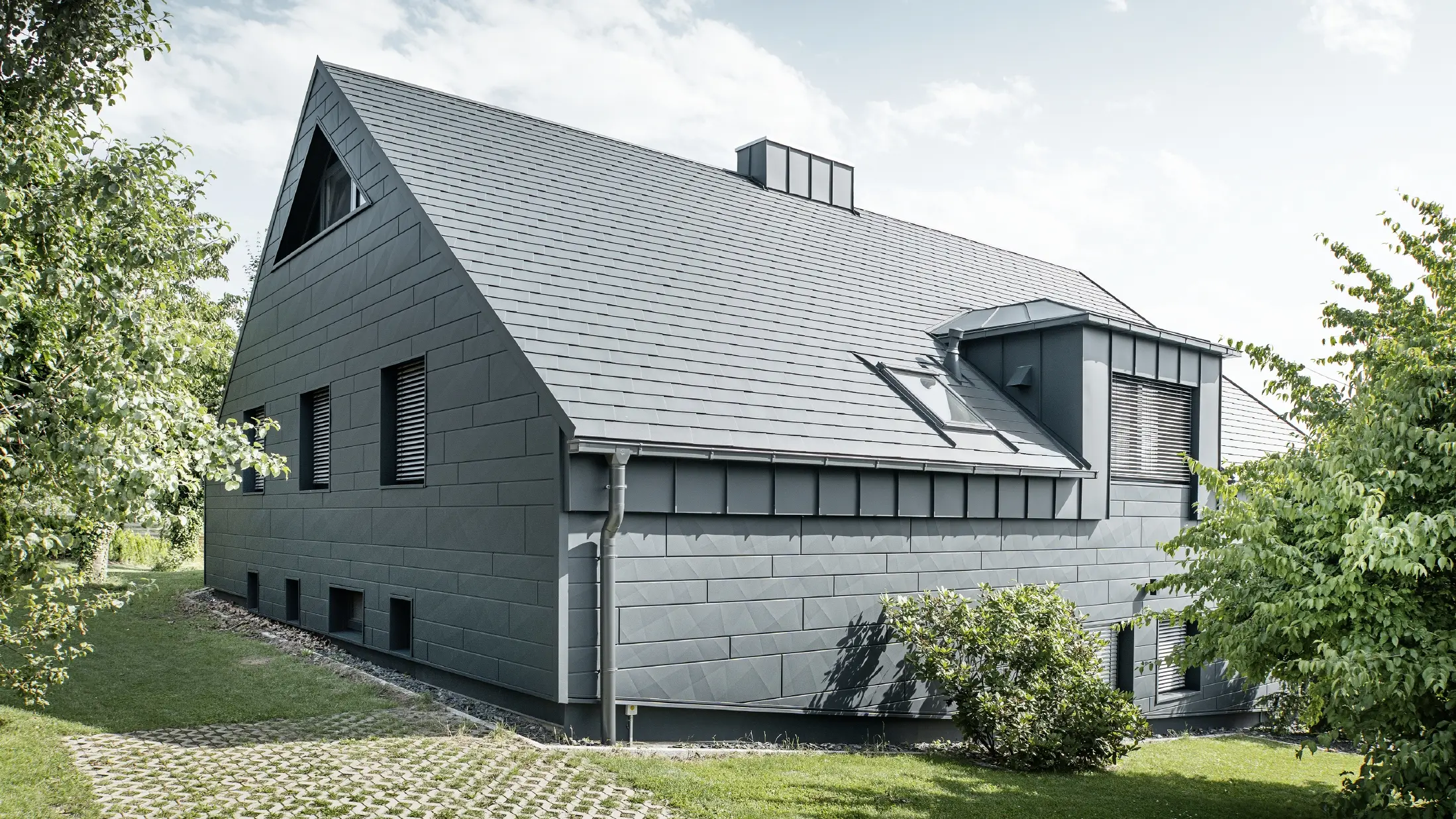 Grande casa unifamiliare con tetto a due falde e molte finestre. L'intera casa è stata rivestita con un involucro in alluminio antracite, sul tetto sono state utilizzate tegole in alluminio PREFA e sulla facciata Doga.X in antracite.