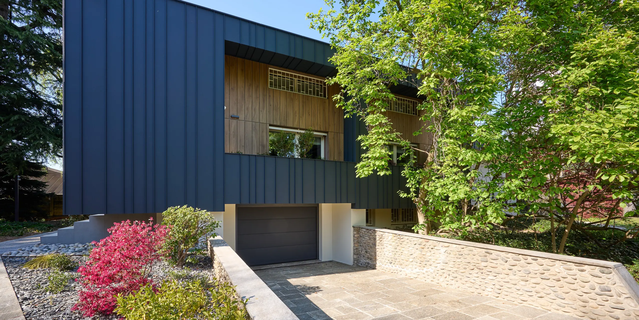 Frontansicht der Villa Castellanza mit eleganter PREFALZ Fassade in P.10 Anthrazit. Die markanten Stehfalzflächen bilden einen spannenden Kontrast zu den warmen Holzelementen und der üppigen Gartenlandschaft. Die hochwertige PREFA Aluminiumhülle überzeugt durch klare Architektur, hohe Witterungsbeständigkeit, Langlebigkeit und ein zeitlos modernes Erscheinungsbild.