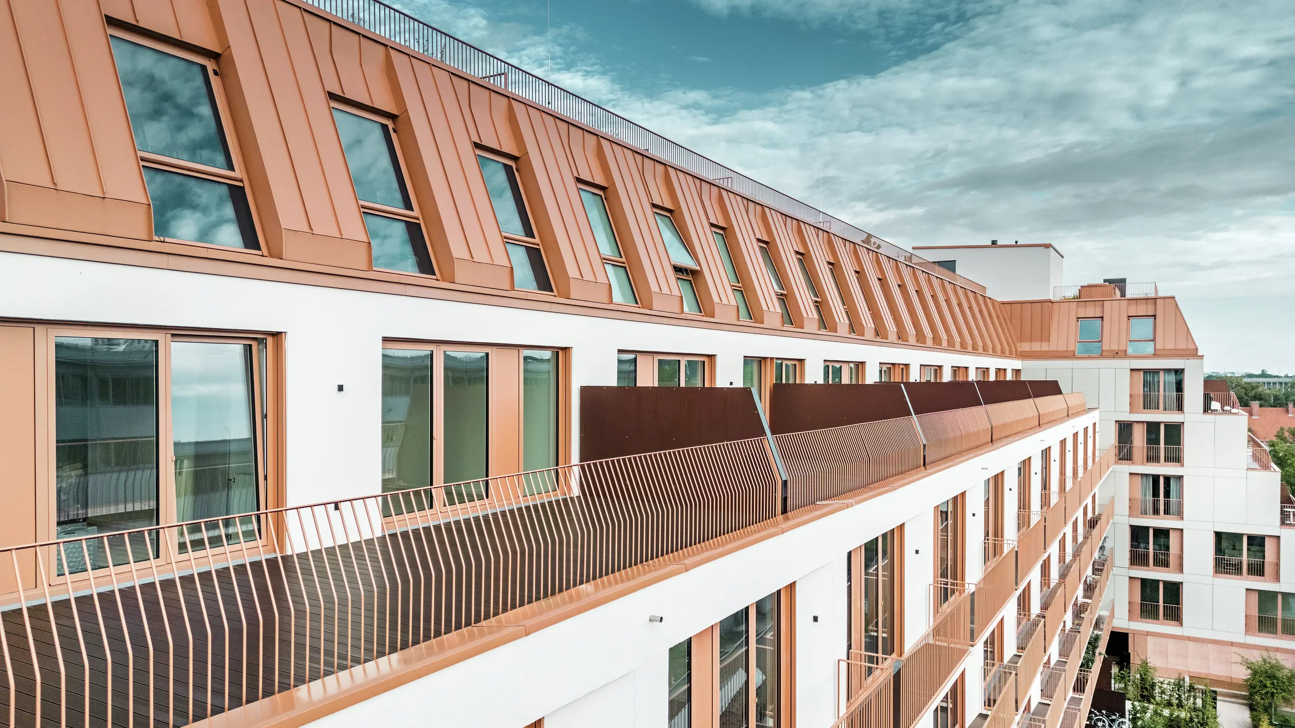 Moderno edificio residenziale Van B a Monaco di Baviera con un suggestivo paesaggio del tetto realizzato in alluminio PREFALZ nel colore speciale IGP Orange HDP.  Il rivestimento del tetto di PREFA, durevole e a bassa manutenzione, garantisce un'unità estetica e un'elevata resistenza agli agenti atmosferici. L'elegante design della facciata, con cornici delle finestre color rame-arancio e generose aree balconate, sottolinea l'innovativo concetto architettonico. La soluzione ideale per progetti di edilizia urbana con elevati standard di design e funzionalità.