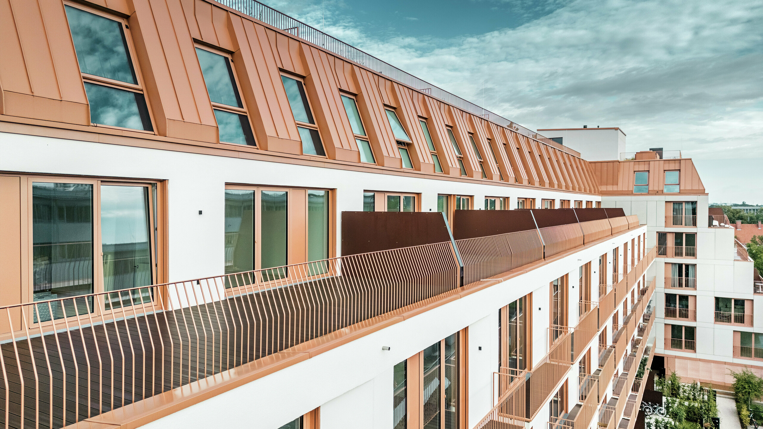 Moderno edificio residenziale Van B a Monaco di Baviera con un suggestivo paesaggio del tetto realizzato in alluminio PREFALZ nel colore speciale IGP Orange HDP.  Il rivestimento del tetto di PREFA, durevole e a bassa manutenzione, garantisce un'unità estetica e un'elevata resistenza agli agenti atmosferici. L'elegante design della facciata, con cornici delle finestre color rame-arancio e generose aree balconate, sottolinea l'innovativo concetto architettonico. La soluzione ideale per progetti di edilizia urbana con elevati standard di design e funzionalità.