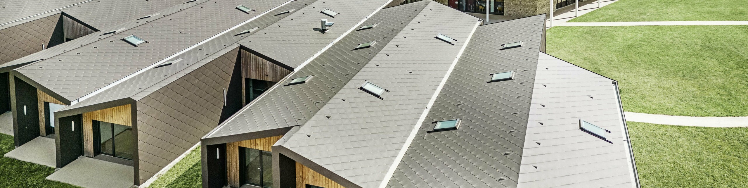 L'immagine mostra una veduta aerea di un moderno complesso edilizio rivestito con scaglie 44 x 44 di PREFA per tetti e facciata, nel colore marrone testa di moro. Il paesaggio del tetto è caratterizzato da forme geometriche combinate con grandi facciate in vetro ed elementi in legno. Sullo sfondo si intravede un'antica chiesa che forma un contrasto armonioso con l'architettura moderna. Il prato verde e la natura circostante sottolineano il design aperto e sostenibile dell'intero complesso.