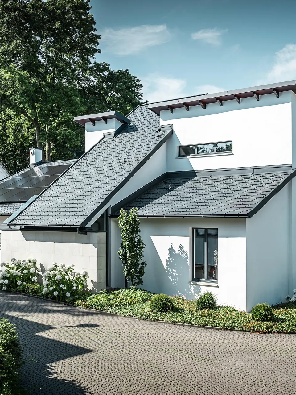 Moderna casa unifamiliare con facciata intonacata di bianco e tetto spiovente PREFA in alluminio scuro. Sul tetto sono visibili moduli fotovoltaici integrati e un sistema di smaltimento acque piovane in alluminio color antracite. L'edificio si trova in un curato complesso residenziale con giardini antistanti, vialetto pavimentato e case unifamiliari circostanti.