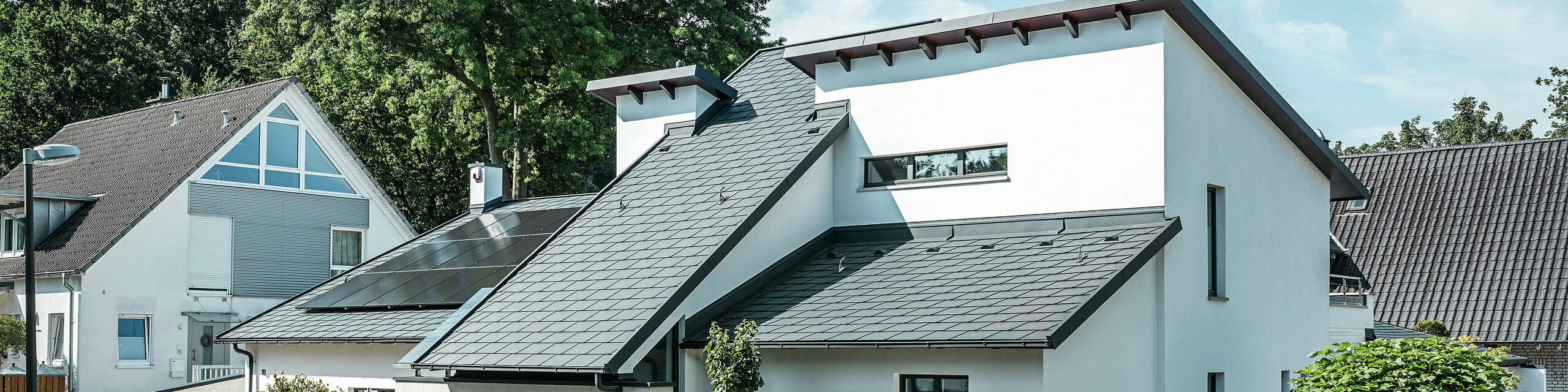 Moderna casa unifamiliare con facciata intonacata di bianco e tetto spiovente PREFA in alluminio scuro. Sul tetto sono visibili moduli fotovoltaici integrati e un sistema di smaltimento acque piovane in alluminio color antracite. L'edificio si trova in un curato complesso residenziale con giardini antistanti, vialetto pavimentato e case unifamiliari circostanti.