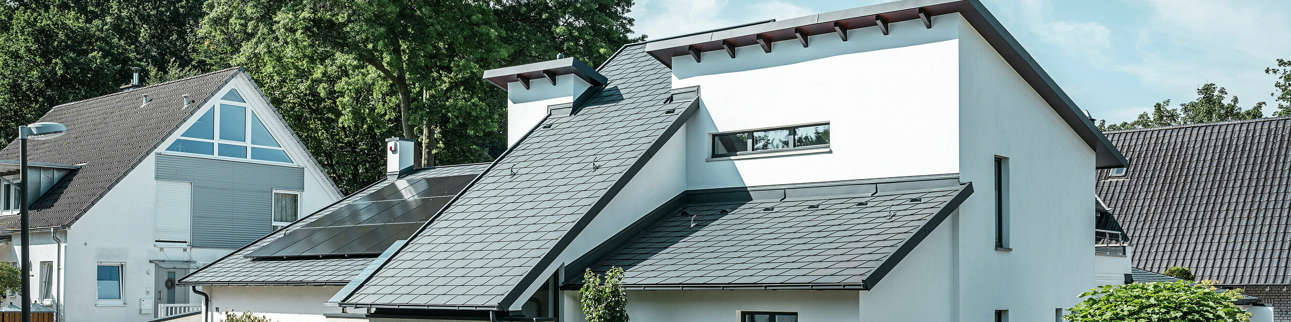 Modernes Einfamilienhaus mit weißer Putzfassade und dunklem PREFA Schrägdach aus Aluminium. Auf dem Dach sind integrierte Photovoltaikmodule und eine Aluminium-Dachentwässerung in Anthrazit zu sehen. Das Gebäude liegt in einer gepflegten Wohnsiedlung mit bepflanzten Vorgärten, gepflasterter Zufahrt und umliegenden Einfamilienhäusern.