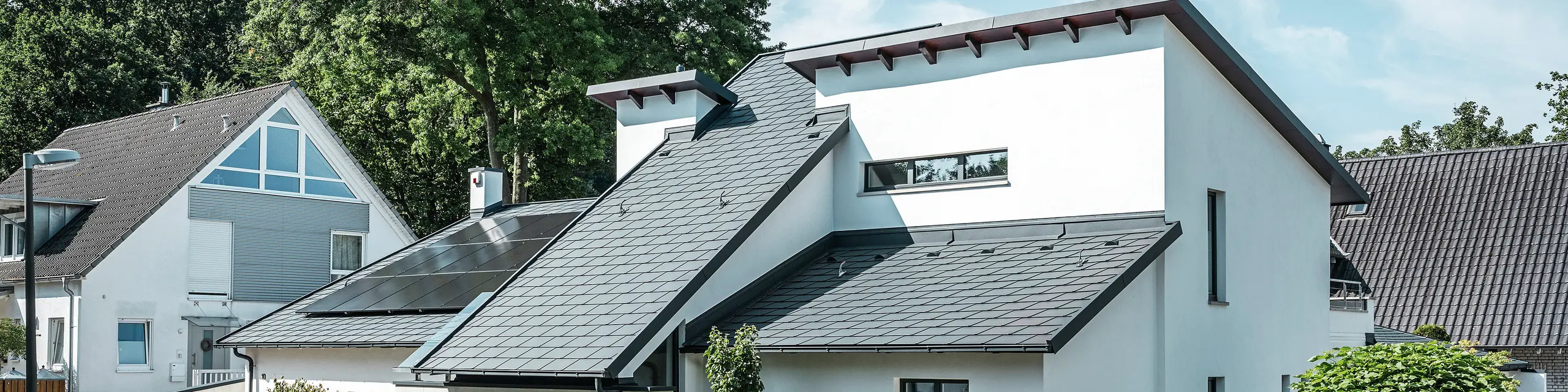 Modernes Einfamilienhaus mit weißer Putzfassade und dunklem PREFA Schrägdach aus Aluminium. Auf dem Dach sind integrierte Photovoltaikmodule und eine Aluminium-Dachentwässerung in Anthrazit zu sehen. Das Gebäude liegt in einer gepflegten Wohnsiedlung mit bepflanzten Vorgärten, gepflasterter Zufahrt und umliegenden Einfamilienhäusern.