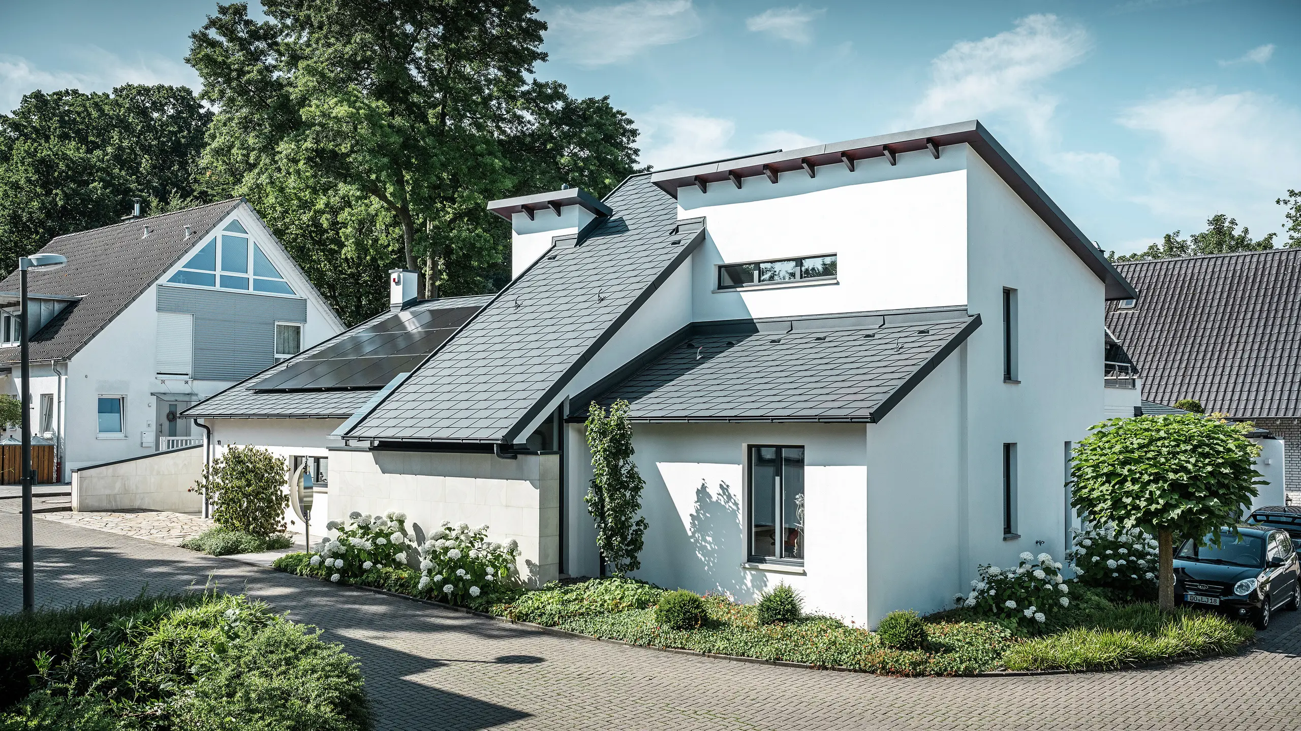 Modernes Einfamilienhaus mit weißer Putzfassade und dunklem PREFA Schrägdach aus Aluminium. Auf dem Dach sind integrierte Photovoltaikmodule und eine Aluminium-Dachentwässerung in Anthrazit zu sehen. Das Gebäude liegt in einer gepflegten Wohnsiedlung mit bepflanzten Vorgärten, gepflasterter Zufahrt und umliegenden Einfamilienhäusern.