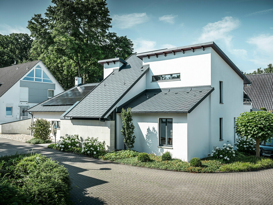 Modernes Einfamilienhaus mit weißer Putzfassade und dunklem PREFA Schrägdach aus Aluminium. Auf dem Dach sind integrierte Photovoltaikmodule und eine Aluminium-Dachentwässerung in Anthrazit zu sehen. Das Gebäude liegt in einer gepflegten Wohnsiedlung mit bepflanzten Vorgärten, gepflasterter Zufahrt und umliegenden Einfamilienhäusern.