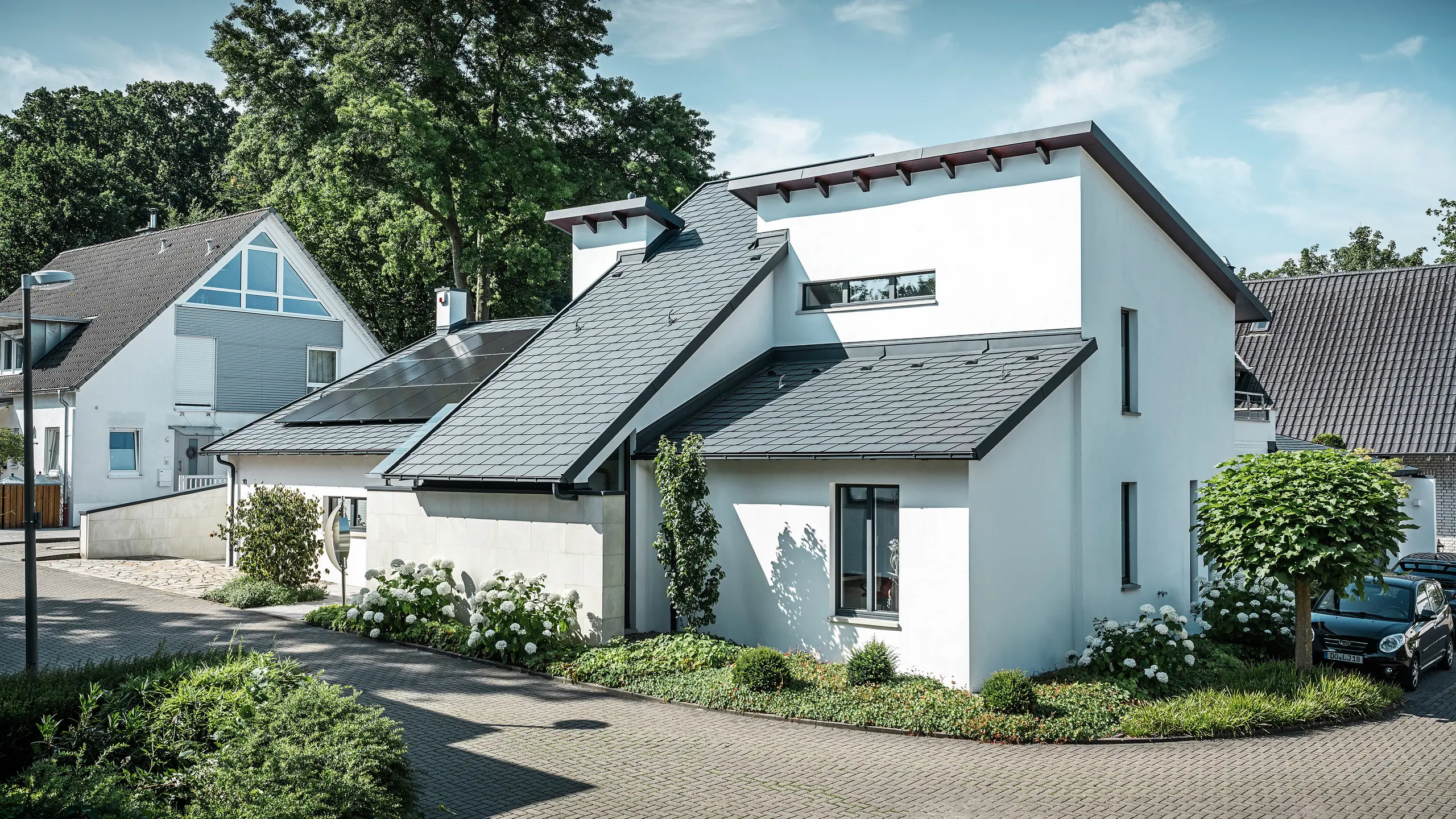 Moderna casa unifamiliare con facciata intonacata di bianco e tetto spiovente PREFA in alluminio scuro. Sul tetto sono visibili moduli fotovoltaici integrati e un sistema di smaltimento acque piovane in alluminio color antracite. L'edificio si trova in un curato complesso residenziale con giardini antistanti, vialetto pavimentato e case unifamiliari circostanti.