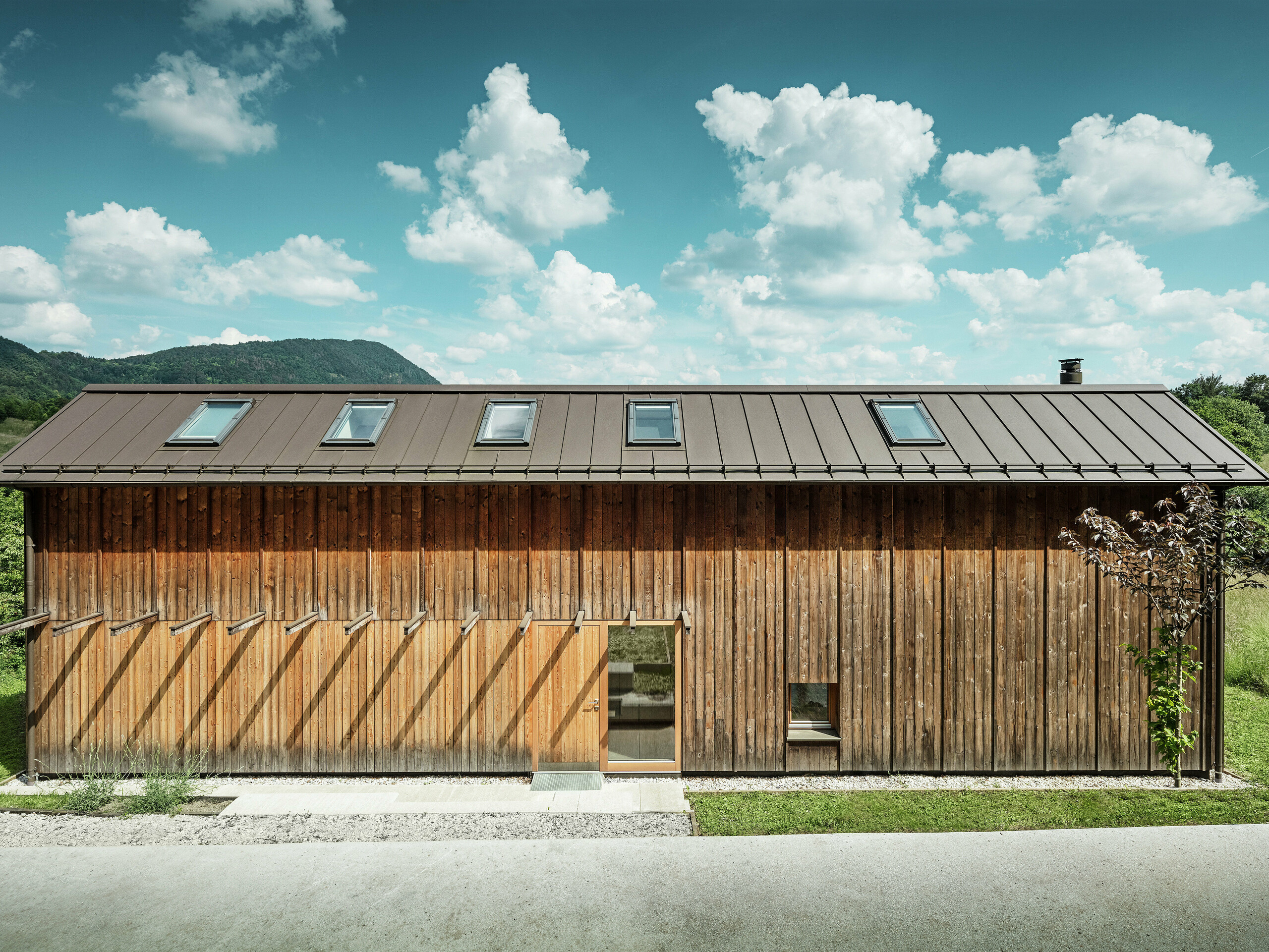 Questa moderna casa sul pendio di una collina colpisce per la sua facciata minimalista in legno e per il resistente tetto in alluminio PREFALZ nel colore P.10 marrone. La copertura del tetto ad aggraffatura verticale convince per l'elevata resistenza agli agenti atmosferici e la durata nel tempo, oltre che per l'armoniosa interazione con la natura circostante. I lucernari integrati e le linee chiare sottolineano l'architettura senza tempo. L'immagine mostra l'area d'ingresso della piccola casa indipendente.