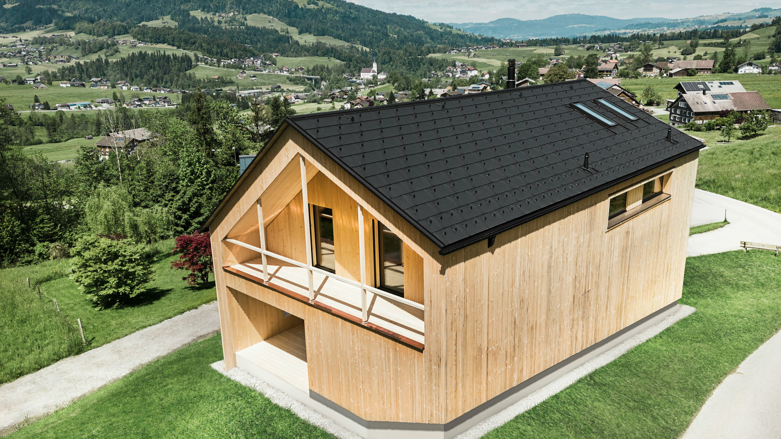 Dieses Foto zeigt ein ökologisches Vorzeigehaus in Egg, das sich mit seiner PREFA Solardachplatte und den schwarzen Dachplatten R.16 gegen die malerische Kulisse eines österreichischen Dorfes abhebt. Die Solarelemente sind nahtlos in die Dachstruktur eingebettet und liefern Energie aus einer erneuerbaren Energiequelle, während das natürliche Holz der Fassade und die sattgrüne Umgebung eine Verbindung von Nachhaltigkeit und Lokalität darstellen. Der Blick auf die umliegende dörfliche Struktur mit traditionellen Bauten im Hintergrund verleiht dem Bild eine zusätzliche Dimension und betont die gelungene Integration moderner, umweltfreundlicher Bauweisen in gewachsene Landschaften.