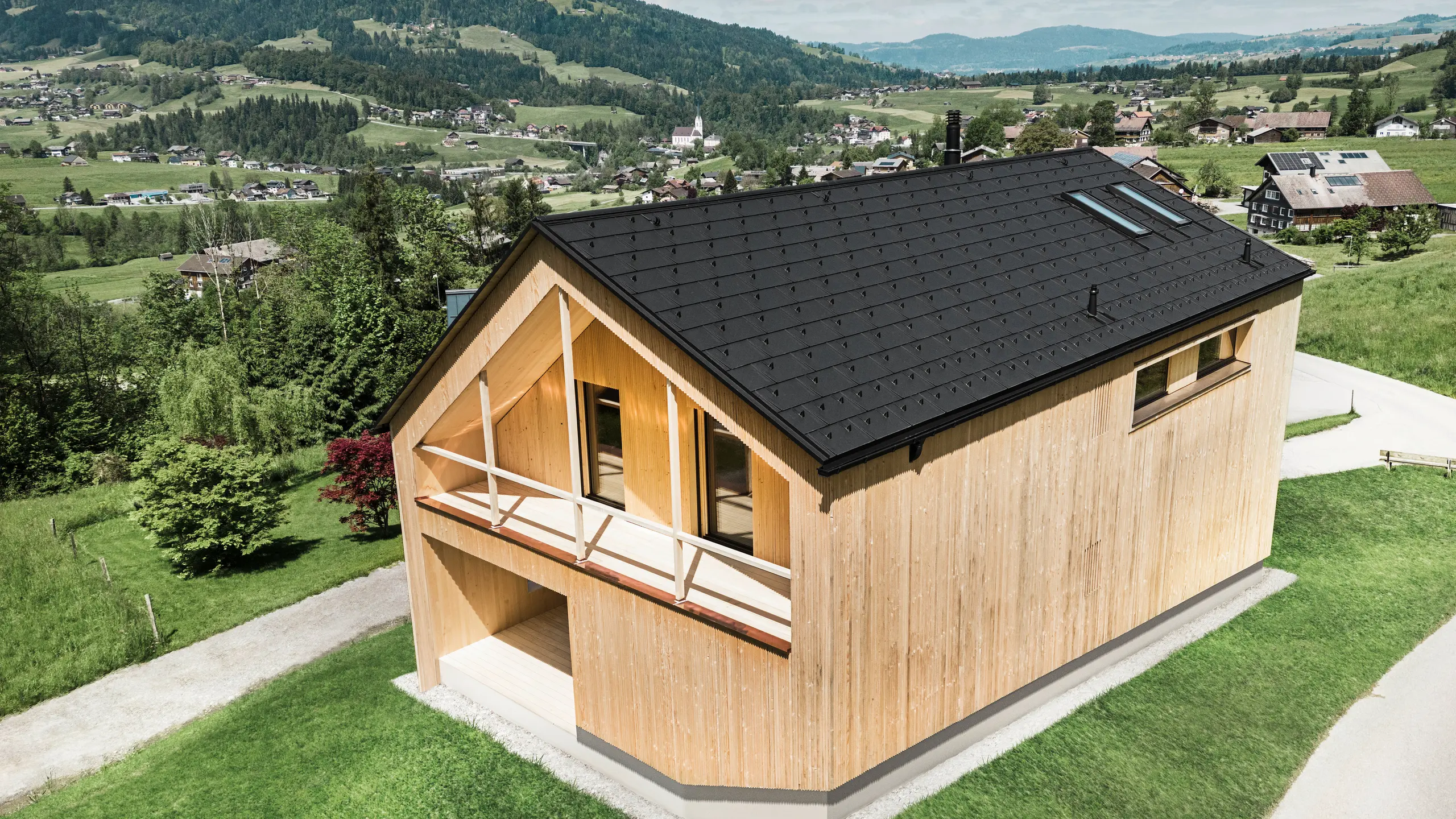 Dieses Foto zeigt ein ökologisches Vorzeigehaus in Egg, das sich mit seiner PREFA Solardachplatte und den schwarzen Dachplatten R.16 gegen die malerische Kulisse eines österreichischen Dorfes abhebt. Die Solarelemente sind nahtlos in die Dachstruktur eingebettet und liefern Energie aus einer erneuerbaren Energiequelle, während das natürliche Holz der Fassade und die sattgrüne Umgebung eine Verbindung von Nachhaltigkeit und Lokalität darstellen. Der Blick auf die umliegende dörfliche Struktur mit traditionellen Bauten im Hintergrund verleiht dem Bild eine zusätzliche Dimension und betont die gelungene Integration moderner, umweltfreundlicher Bauweisen in gewachsene Landschaften.