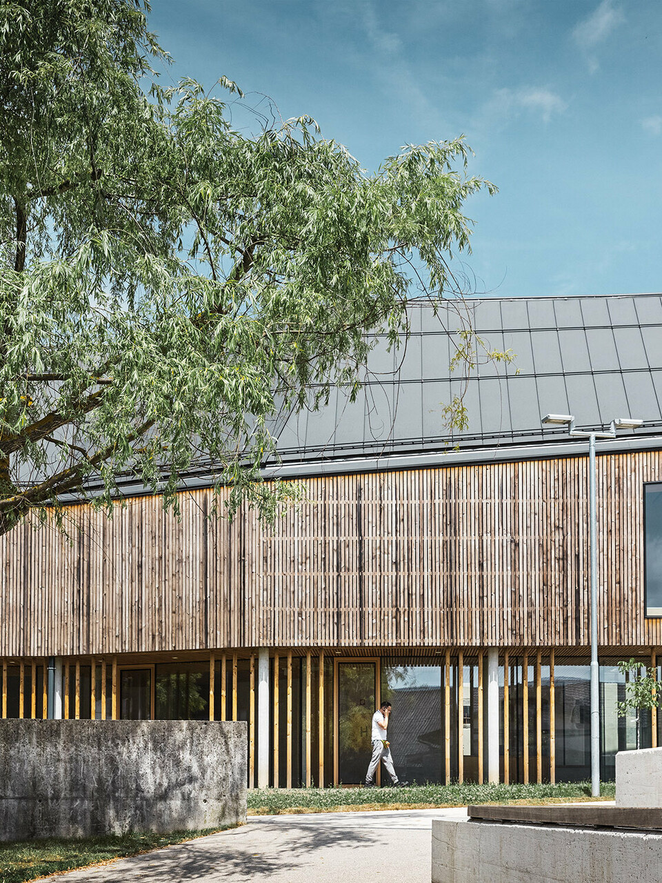 Das neue Pfahlbautenmuseum mit seiner Holzlamellenfassade und dem komplexen, dunkelgrauen PREFA Dach von der Seite; ein Baum rahmt den Bau links oben ein.