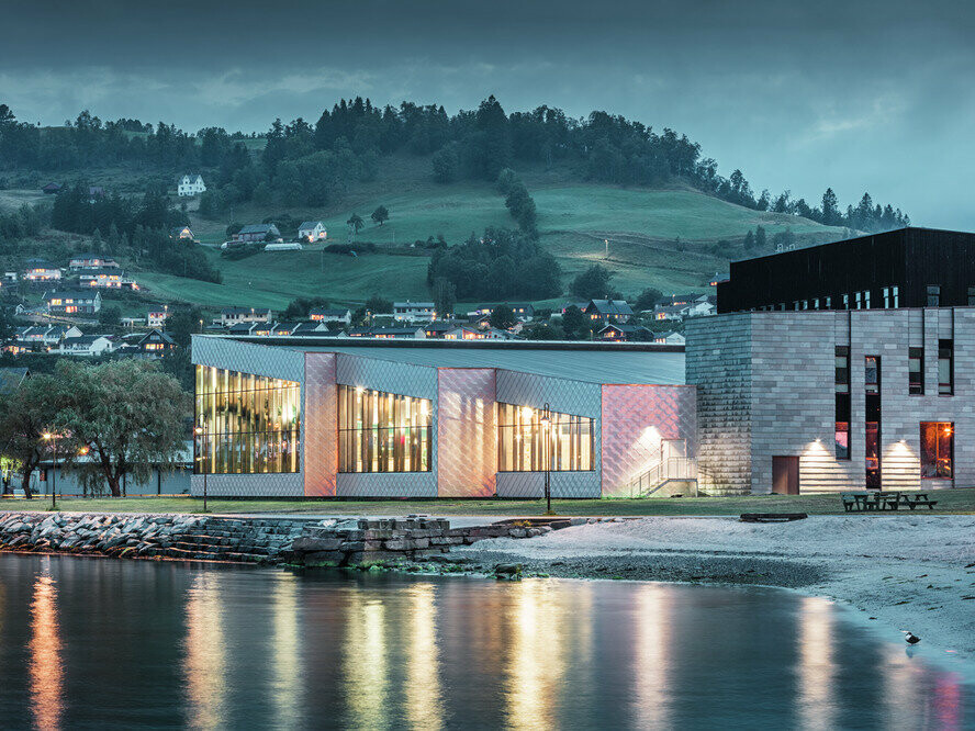 Nella foto, l'edificio polifunzionale dell'Hardangerbadet Healthcare Center di Oystese visto dall'acqua. Rivestito in Scaglia 44 x 44 colore alluminio naturale. Si vede il progetto illuminato all'imbrunire e sullo sfondo Oystese.