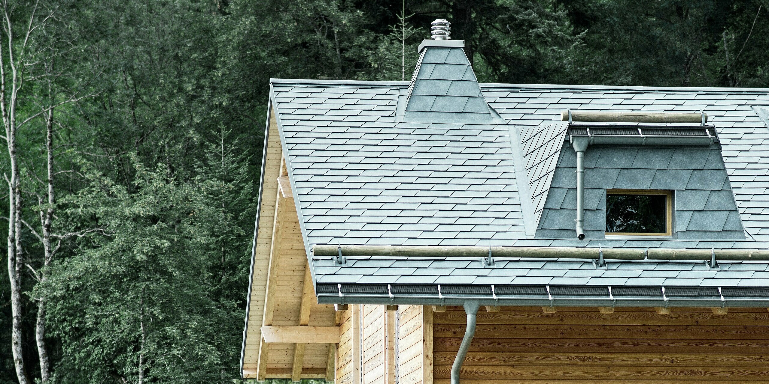 Seitenansicht einer Holz-Lodge am Genfersee mit PREFA Aluminium-Dachschindeln in Steingrau. Die Dachstruktur mit dem markanten Kamin und dem Schneefang aus Rundholz fügt sich harmonisch in die Umgebung ein. Das langlebige und ästhetische Blechdach bietet nicht nur Schutz vor den Elementen, sondern stellt auch eine stilvolle Ergänzung zur Holzfassade dar.