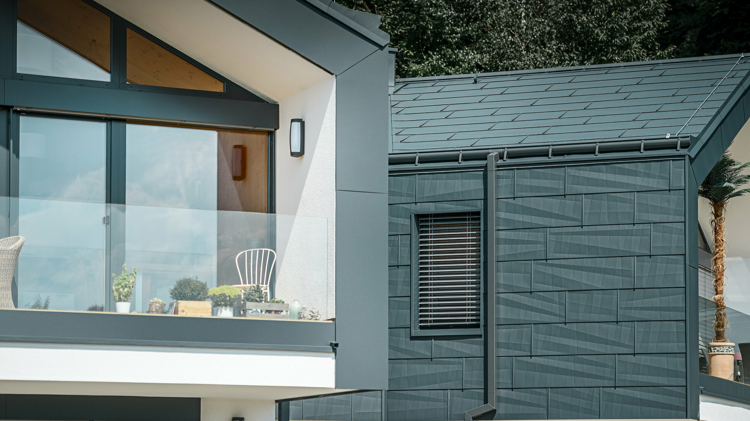 Das Bild zeigt ein modernes Doppelhaus mit einer stilvollen Fassade aus Aluminium. Die Fassade ist in einem dunklen Anthrazitton gehalten und hat eine strukturierte Oberfläche durch das PREFA Dach- und Fassadenpaneel FX.12. Am Dach wurde eine Saumrinne und das Quadratrohr als Ablaufrohr von PREFA montiert. Ein Balkon mit Glasgeländer ist an der oberen Etage zu sehen, während an der unteren Etage Fenster mit Lamellenjalousien zu finden sind. Die Architektur wirkt klar und minimalistisch, und im Hintergrund sind Bäume zu erkennen, die eine natürliche Atmosphäre schaffen.