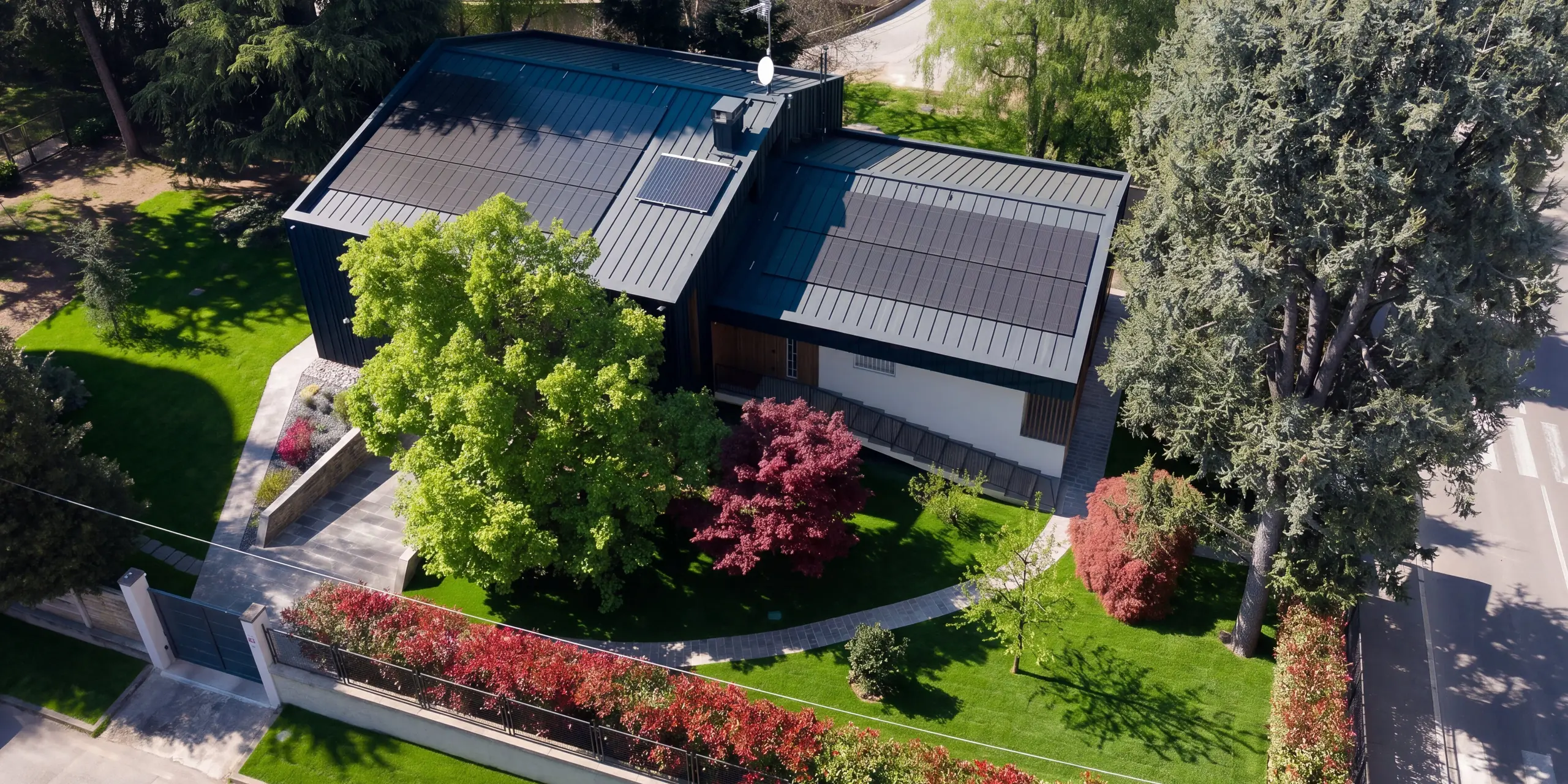 Spektakuläre Luftaufnahme der Villa Castellanza mit PREFALZ Dach und Fassade in P.10 Anthrazit. Großflächig integrierte PREFALZ Solarmodule prägen die moderne Gebäudehülle und ermöglichen nachhaltige Energiegewinnung. Eingebettet in einen gepflegten Garten und alten Baumbestand zeigt das Wohnhaus die perfekte Verbindung aus PREFA Aluminium, Design und Funktion.