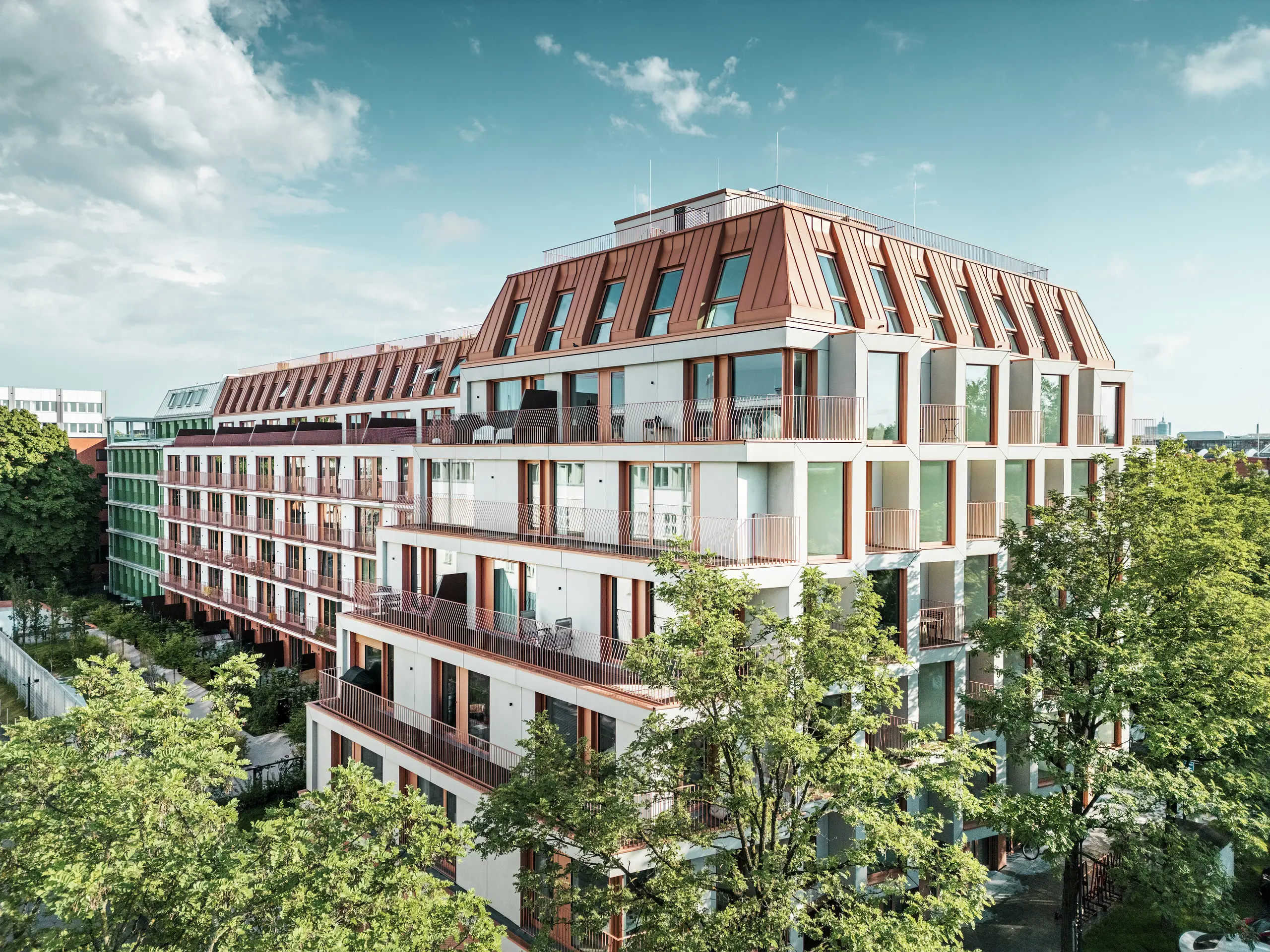 Vista generale del progetto residenziale urbano Van B a Monaco di Baviera, caratterizzato da un'architettura chiara, aree verdi esterne e dal sorprendente tetto in alluminio PREFALZ in IGP Orange HDP di PREFA.  Il materiale del tetto, durevole e a bassa manutenzione, costituisce l'elemento di design unificante dell'edificio e fornisce una soluzione estetica e duratura nello spazio urbano.Le ampie superfici finestrate, i balconi e le planimetrie ben studiate offrono un comfort abitativo moderno con un'elevata qualità del design.