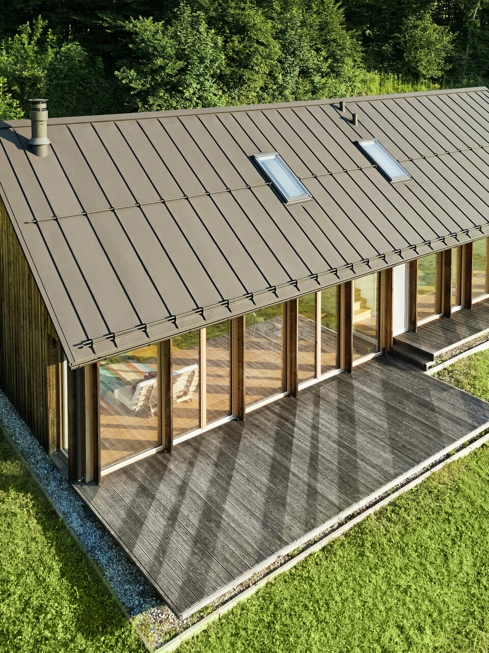 Moderna casa in legno con generosa facciata in vetro e resistente tetto in alluminio PREFALZ in P.10 marrone - ideale per siti in pendenza come Vinje. La copertura del tetto di alta qualità garantisce la massima resistenza agli agenti atmosferici, una lunga durata e un aspetto elegante. L'installazione precisa delle giunture verticali e i lucernari integrati creano un concetto abitativo sostenibile e inondato di luce. La vista a volo d'uccello mostra la precisa lavorazione delle aggraffature in alluminio.