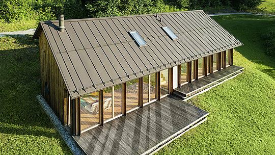 Moderna casa in legno con generosa facciata in vetro e resistente tetto in alluminio PREFALZ in P.10 marrone - ideale per siti in pendenza come Vinje. La copertura del tetto di alta qualità garantisce la massima resistenza agli agenti atmosferici, una lunga durata e un aspetto elegante. L'installazione precisa delle giunture verticali e i lucernari integrati creano un concetto abitativo sostenibile e inondato di luce. La vista a volo d'uccello mostra la precisa lavorazione delle aggraffature in alluminio.