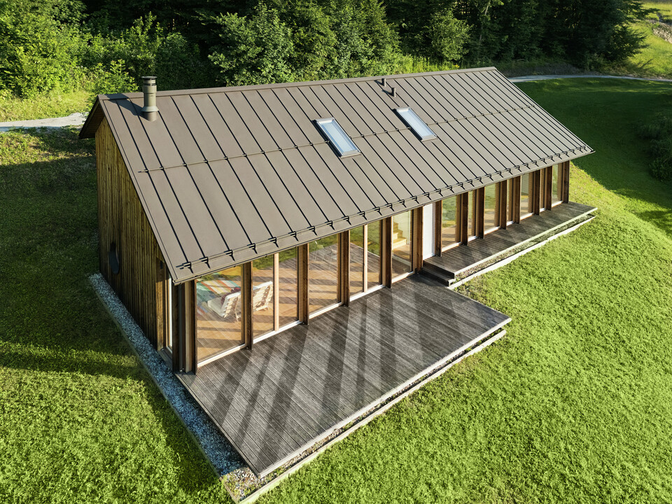 Moderna casa in legno con generosa facciata in vetro e resistente tetto in alluminio PREFALZ in P.10 marrone - ideale per siti in pendenza come Vinje. La copertura del tetto di alta qualità garantisce la massima resistenza agli agenti atmosferici, una lunga durata e un aspetto elegante. L'installazione precisa delle giunture verticali e i lucernari integrati creano un concetto abitativo sostenibile e inondato di luce. La vista a volo d'uccello mostra la precisa lavorazione delle aggraffature in alluminio.
