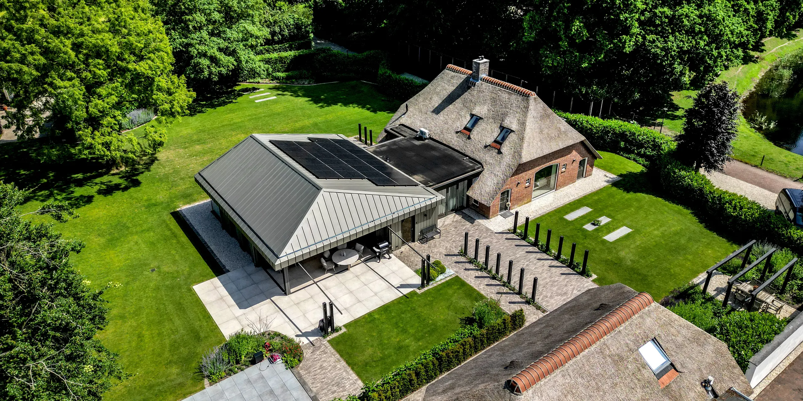 Beeindruckende Luftaufnahme des Einfamilienhauses in Putten mit modernem PREFALZ Anbau und historischem Reetdach. Die markante Aluminiumhülle in P.10 Prefabronze, die präzisen Stehfalze und die integrierten Solarmodule setzen starke architektonische Akzente. Umgeben von einer weitläufigen Parklandschaft zeigt PREFA Aluminium seine Stärken: langlebig, witterungsbeständig, nachhaltig und formschön.