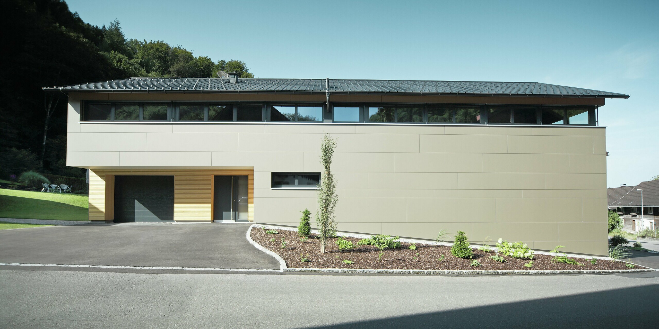 Das Einfamilienhaus in Hohenems präsentiert sich in einer modernen und stilvollen Fassade aus PREFA Aluminium Verbundplatten in Bronzemetallic, mit einem darüberliegenden Dach aus PREFA Dachplatten in P.10 Anthrazit. Die klare Linienführung und das durchdachte Farbkonzept des Designs integrieren das Gebäude nahtlos in die natürliche Umgebung. Die Gestaltung zeigt die Kombination aus Funktionalität und Ästhetik, die PREFA Produkte für zeitgemäße Wohnarchitektur bieten.