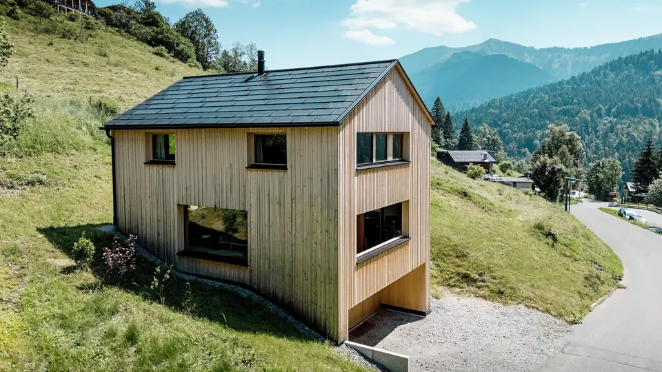 In alto sopra la valle, la casa Immi si erge come un monolite di legno dalle linee pulite sul soleggiato versante montuoso di Ebnit. La facciata essenziale in abete bianco e il tetto solare nero PREFA creano un forte contrasto con l’ampio paesaggio alpino. Le tegole fotovoltaiche integrate nel tetto e le tegole R.16 in P.10 nero forniscono energia elettrica, resistono alle intemperie alpine e garantiscono un aspetto armonioso e duraturo del tetto.