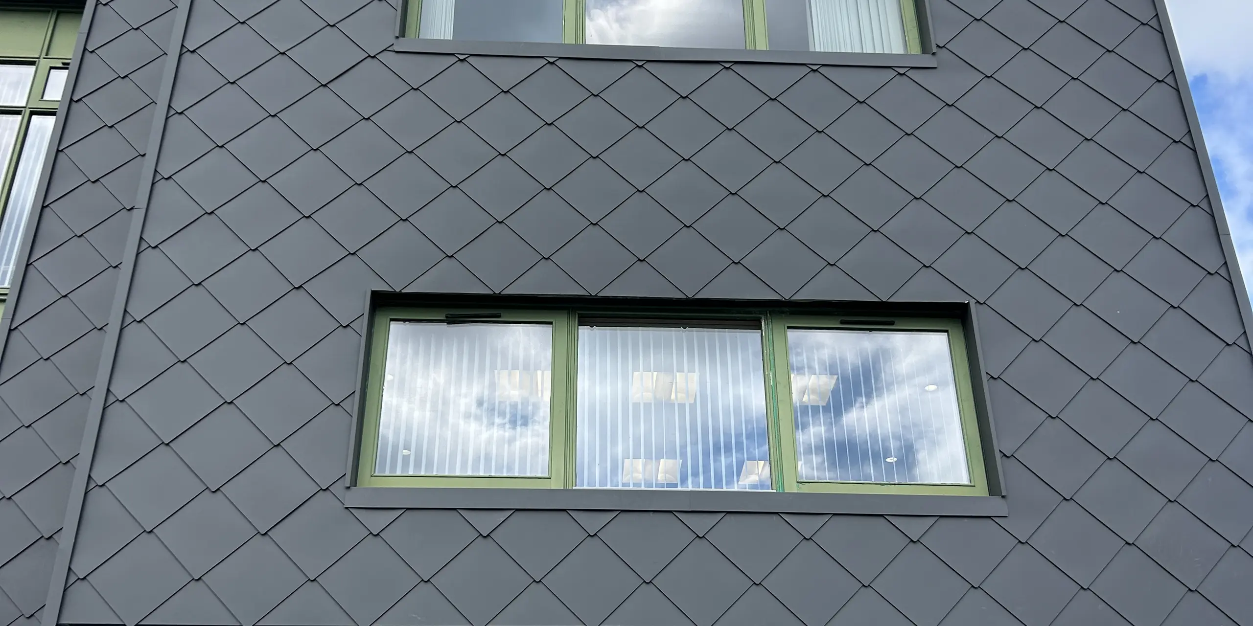 Dynamische Nahaufnahme der mit PREFA Wandrauten 44 x 44 in der Farbe P.10 Dunkelgrau verkleideten Fassade des Zentrums für psychische Gesundheit von Kindern und Jugendlichen in Wexford: Die präzise Rautenstruktur erzeugt lebendige Licht- und Schattenspiele, während die grün gerahmten Fenster Kontraste setzen. Die robuste Aluminiumfassade überzeugt durch Langlebigkeit, Wartungsarmut und hohe Witterungsbeständigkeit.